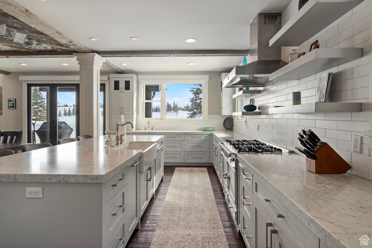 Kitchen featuring tasteful backsplash, beam ceiling, wall chimney exhaust hood, a kitchen island with sink, and white cabinetry