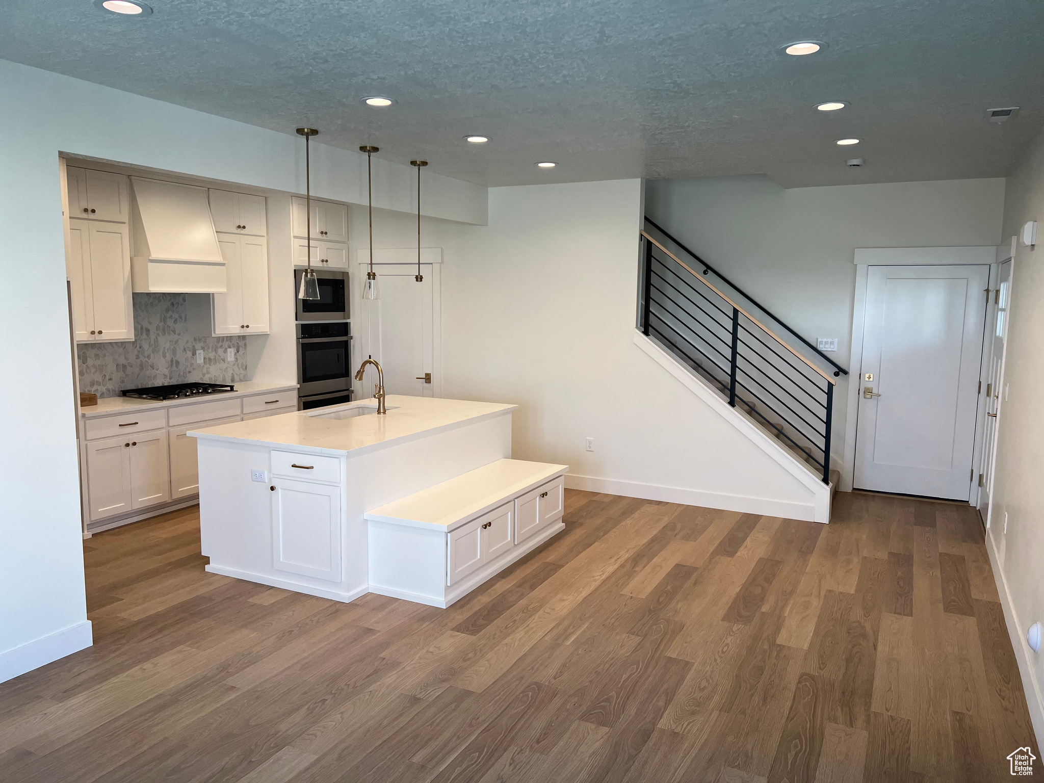 Kitchen with custom range hood, appliances with stainless steel finishes, wood-type flooring, decorative light fixtures, and a kitchen island with sink