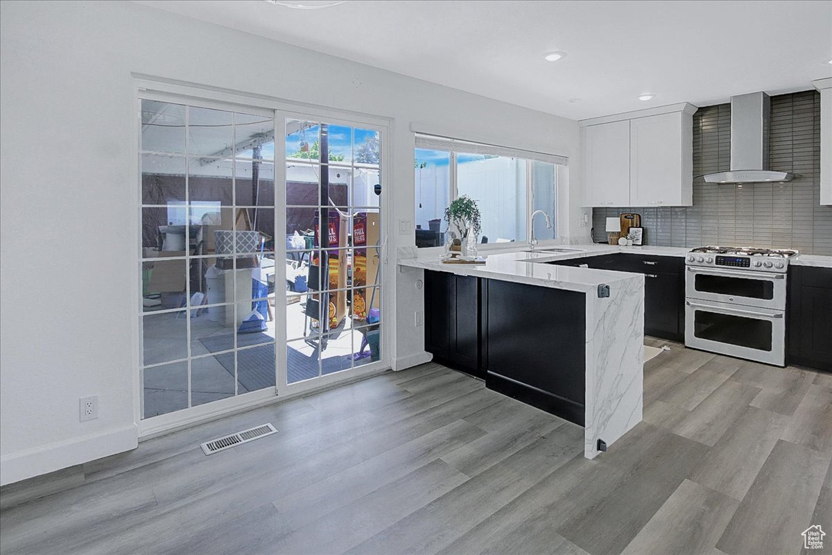 Kitchen featuring light hardwood / wood-style flooring, tasteful backsplash, range with two ovens, wall chimney range hood, and white cabinets