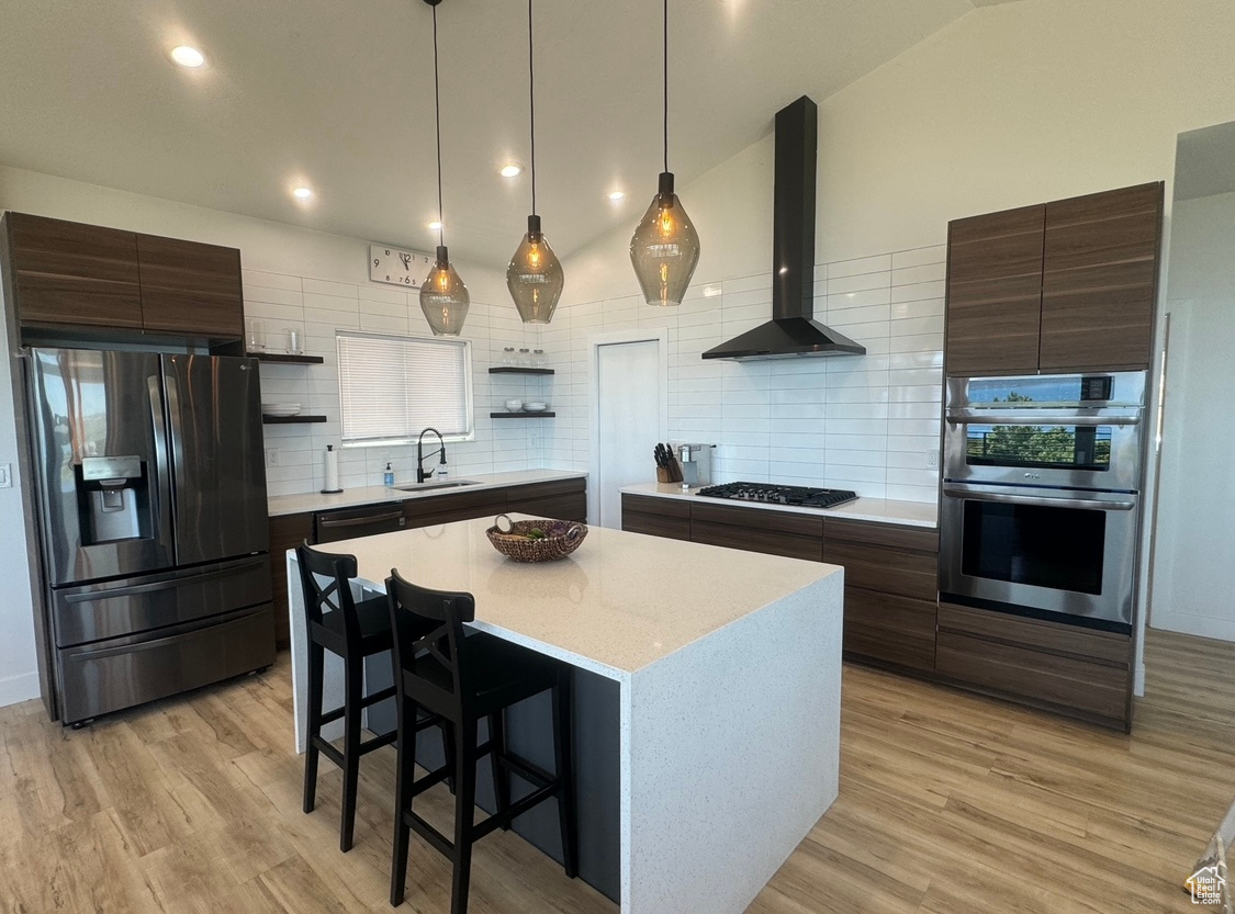 Kitchen with wall chimney range hood, light hardwood / wood-style floors, a kitchen island, backsplash, and stainless steel appliances