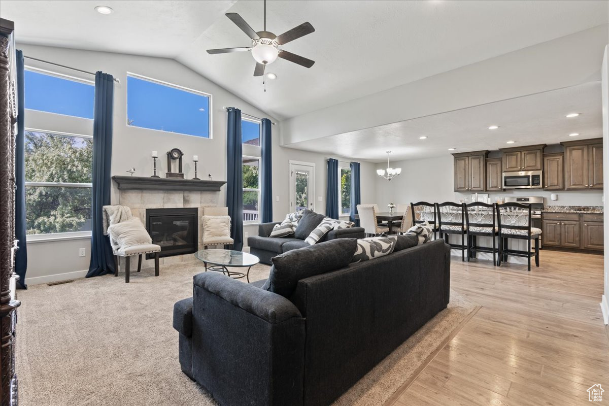 Living room with light wood-type flooring, a tiled fireplace, lofted ceiling, and ceiling fan with notable chandelier