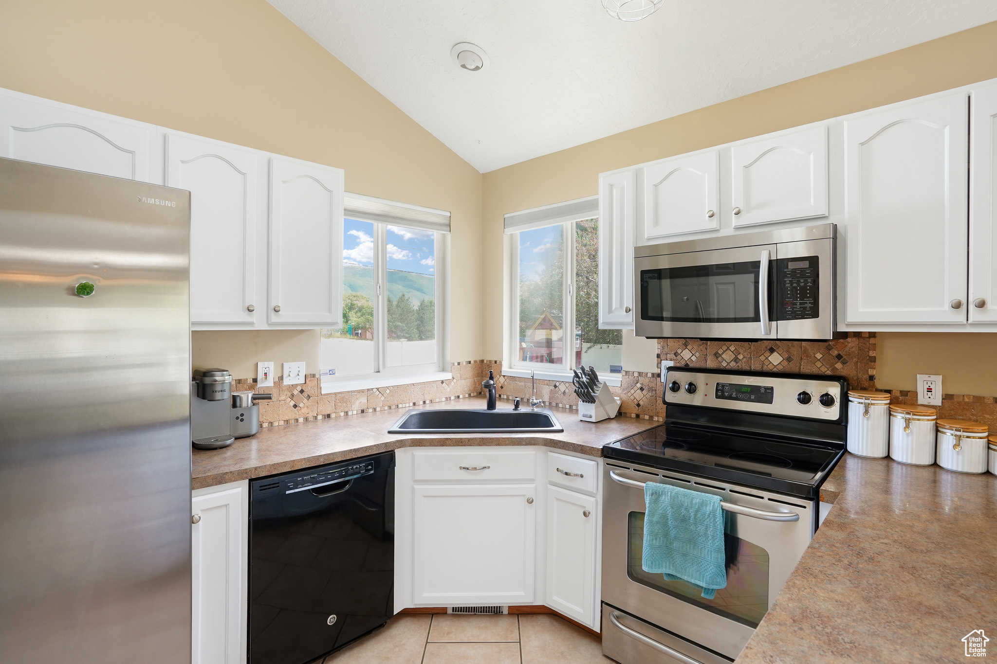 Kitchen with stainless steel appliances, white cabinetry, sink, light tile floors, and lofted ceiling