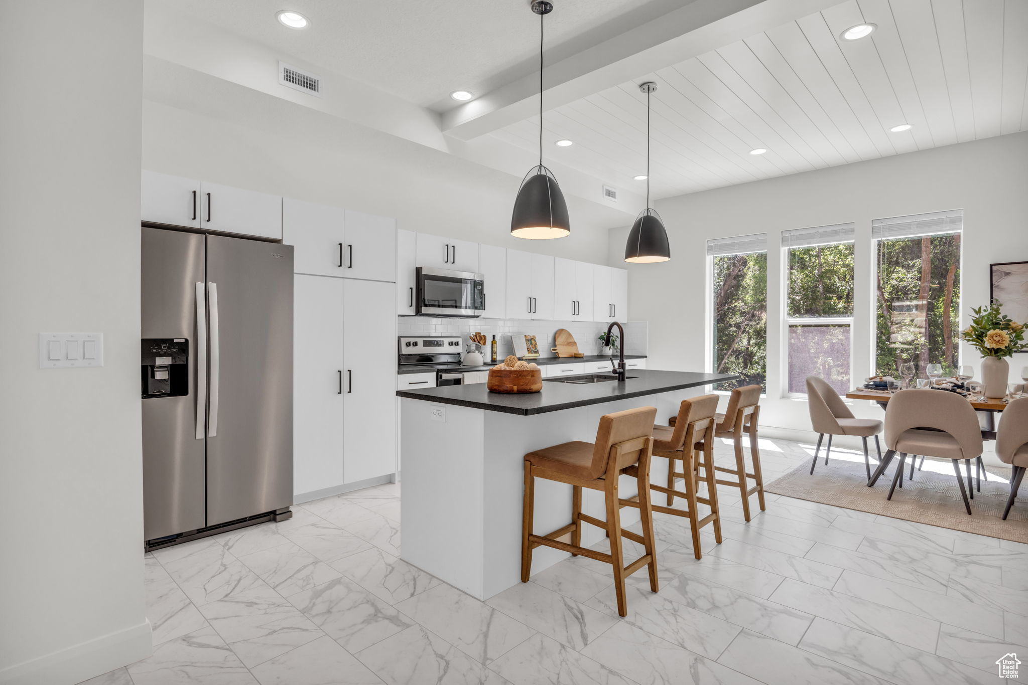 Kitchen featuring beamed ceiling, light tile floors, a kitchen island with sink, white cabinetry, and appliances with stainless steel finishes