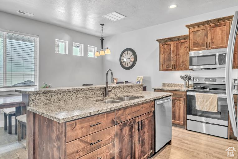 Kitchen featuring hanging light fixtures, appliances with stainless steel finishes, light hardwood / wood-style floors,  and a kitchen island with sink