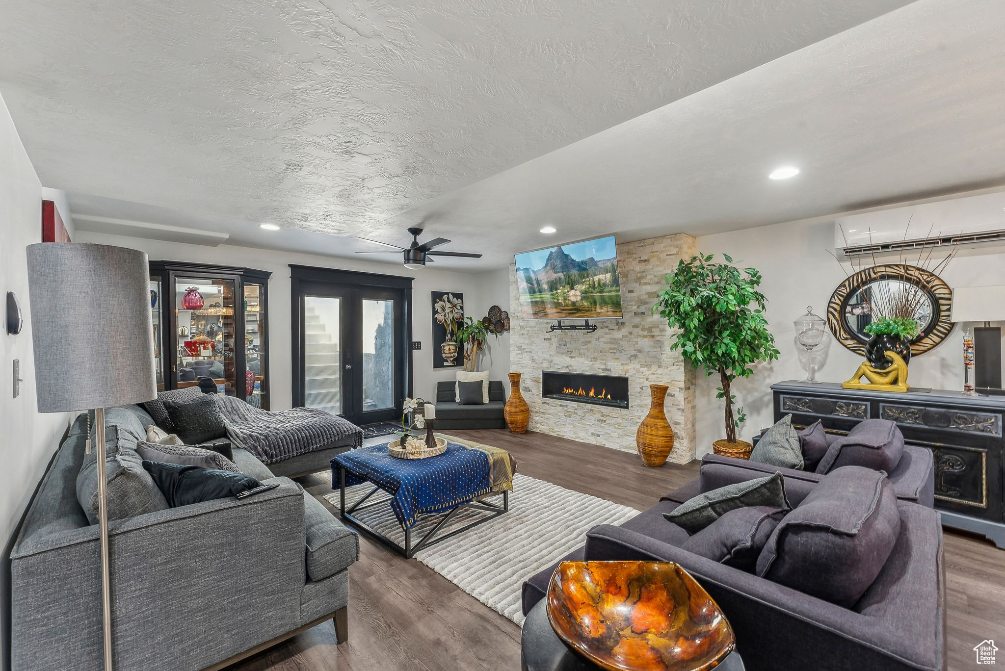 Living room with a stone fireplace, an AC wall unit, wood-type flooring, and ceiling fan