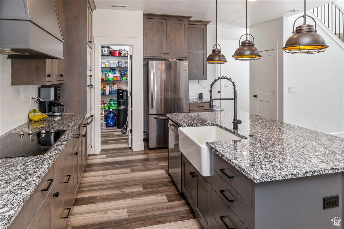 Kitchen with an island with sink, stainless steel appliances, custom exhaust hood, decorative backsplash, and hardwood / wood-style flooring