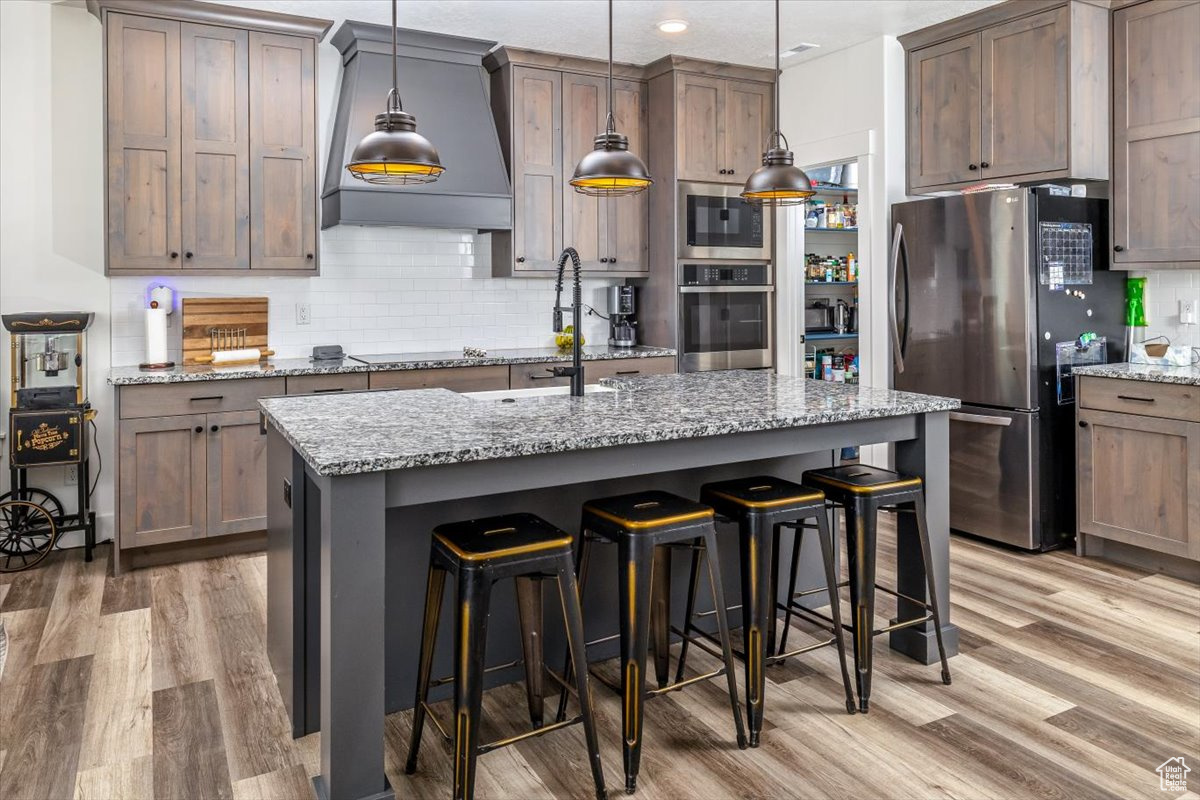 Kitchen with decorative light fixtures, an island with sink, custom range hood, light hardwood / wood-style floors, and appliances with stainless steel finishes