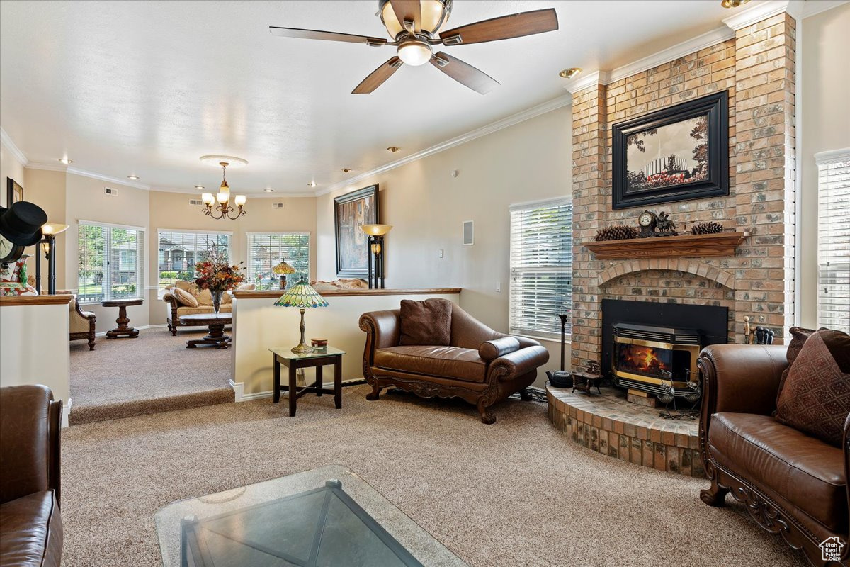 Carpeted living room featuring a fireplace, ceiling fan with notable chandelier, brick wall, and ornamental molding