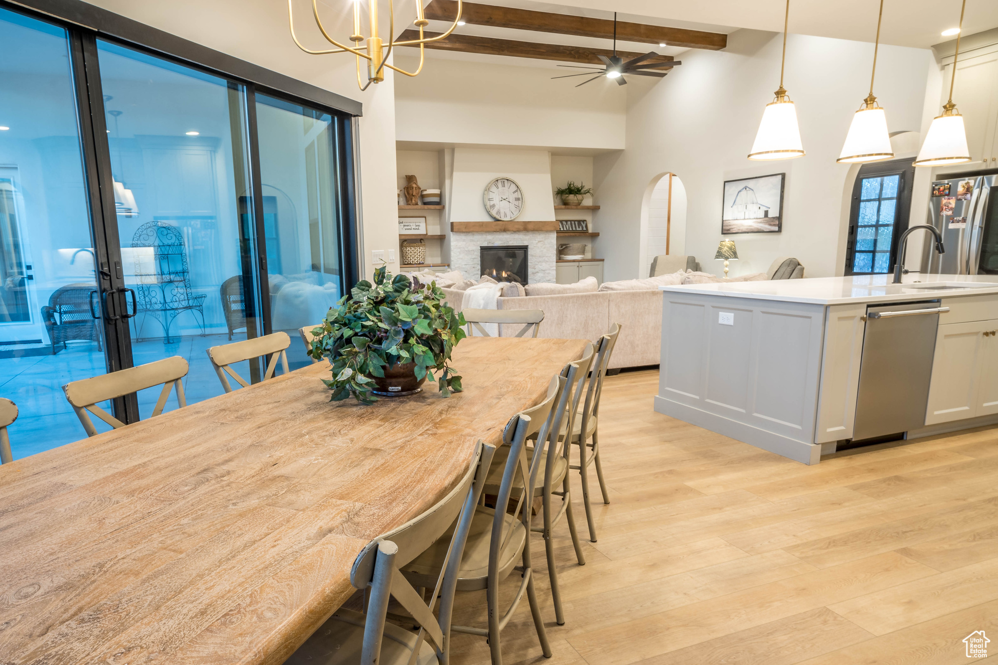 Dining room with light hardwood / wood-style flooring, ceiling fan with notable chandelier, a stone fireplace, beamed