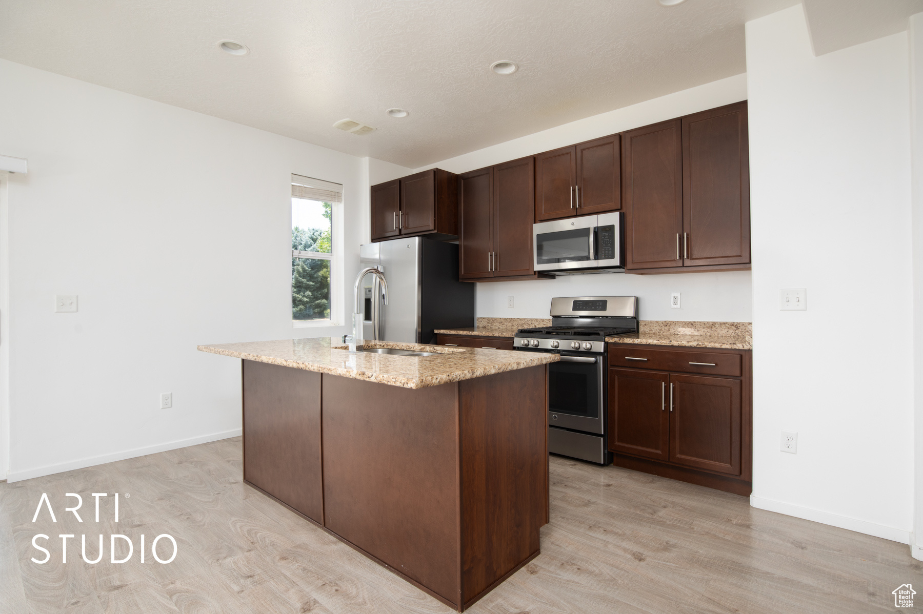 Kitchen featuring light wood-type flooring, light stone countertops, dark brown cabinetry, appliances with stainless steel finishes, and a kitchen island with sink