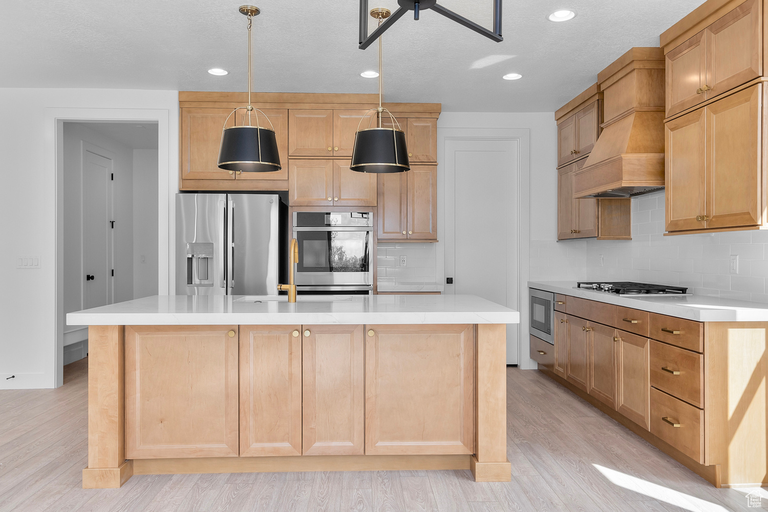 Kitchen featuring decorative light fixtures, a center island with sink, appliances with stainless steel finishes, custom exhaust hood, and light wood-type flooring