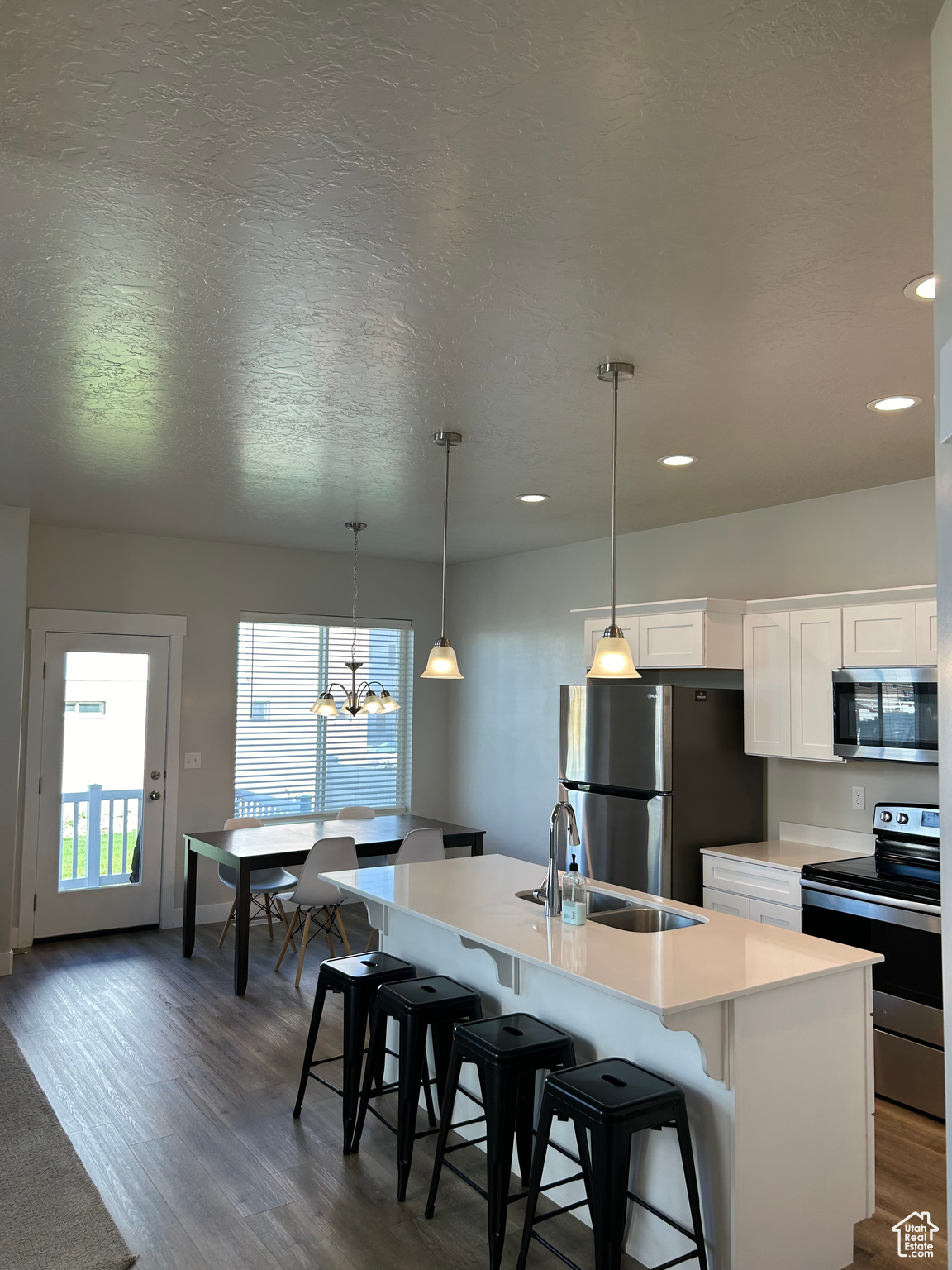 Kitchen featuring wood-style flooring, decorative light fixtures, stainless steel appliances, a center island with sink, and white cabinets.