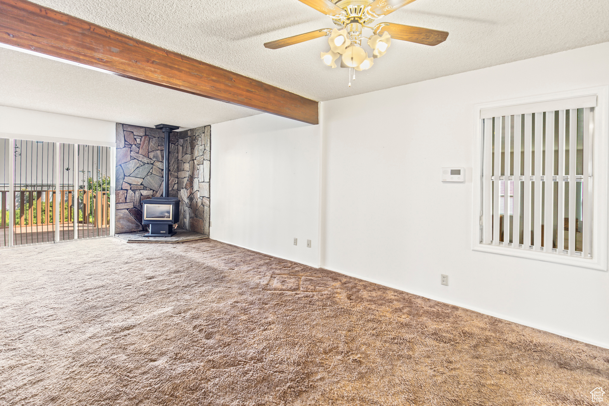 Unfurnished living room featuring carpet flooring, ceiling fan, a wood stove, and beam ceiling