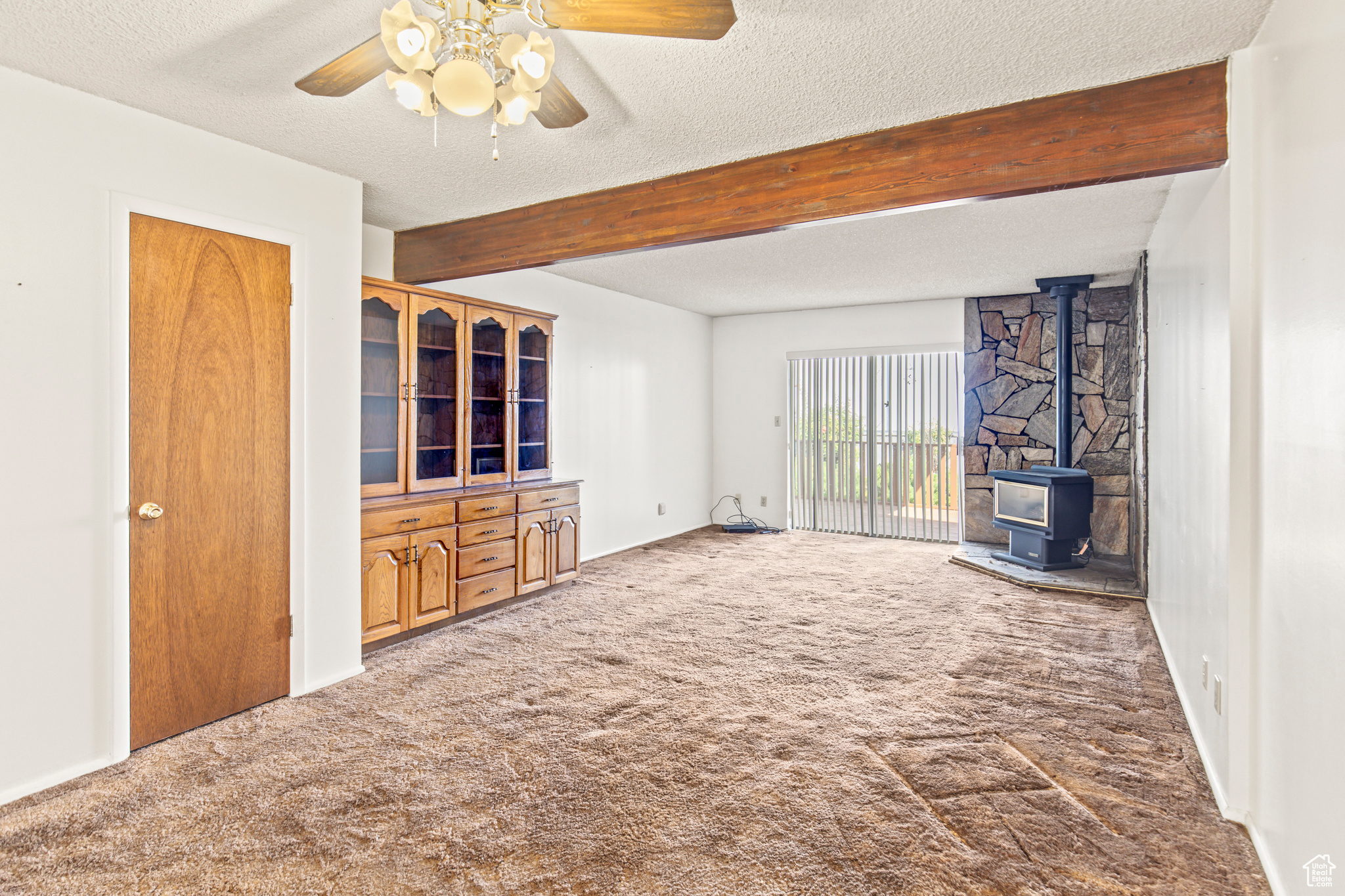 Unfurnished living room featuring a wood stove, carpet, ceiling fan, beam ceiling, and a textured ceiling