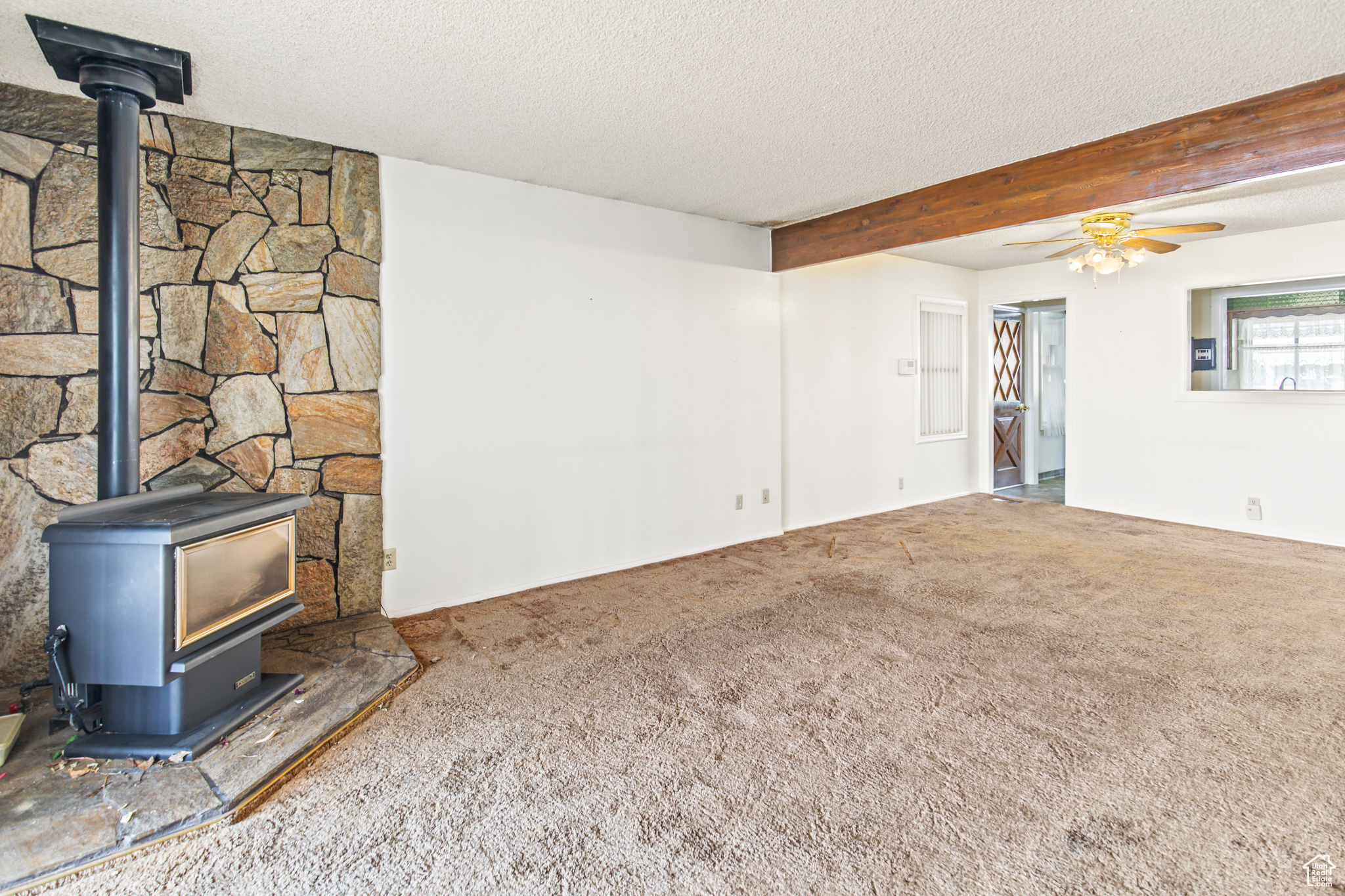 Unfurnished living room featuring ceiling fan, a textured ceiling, beam ceiling, and a wood stove