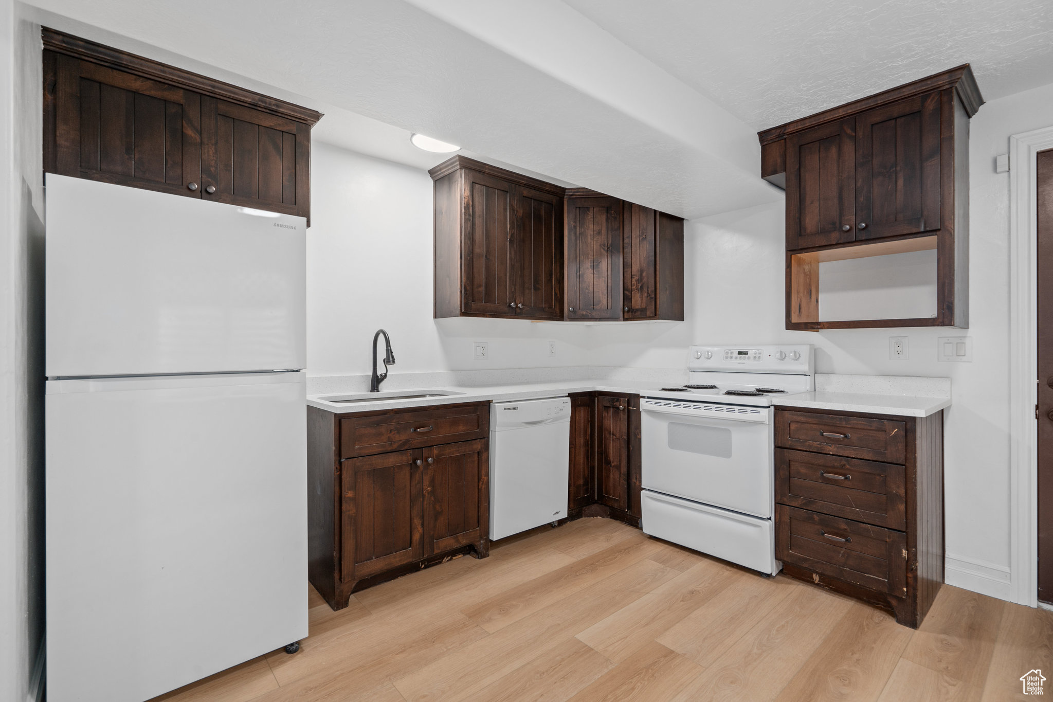 Kitchen with dark brown cabinetry, sink, light hardwood / wood-style floors, and white appliances