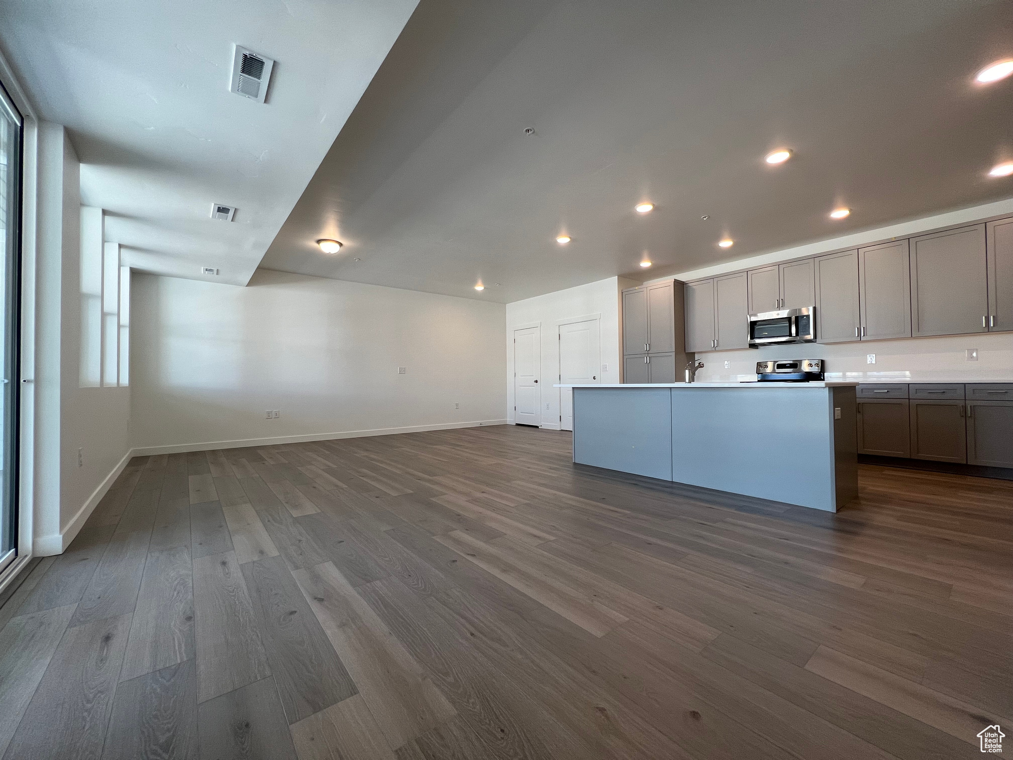 Kitchen with gray cabinets, dark wood-type flooring, stainless steel appliances, a center island with sink, and a healthy amount of sunlight