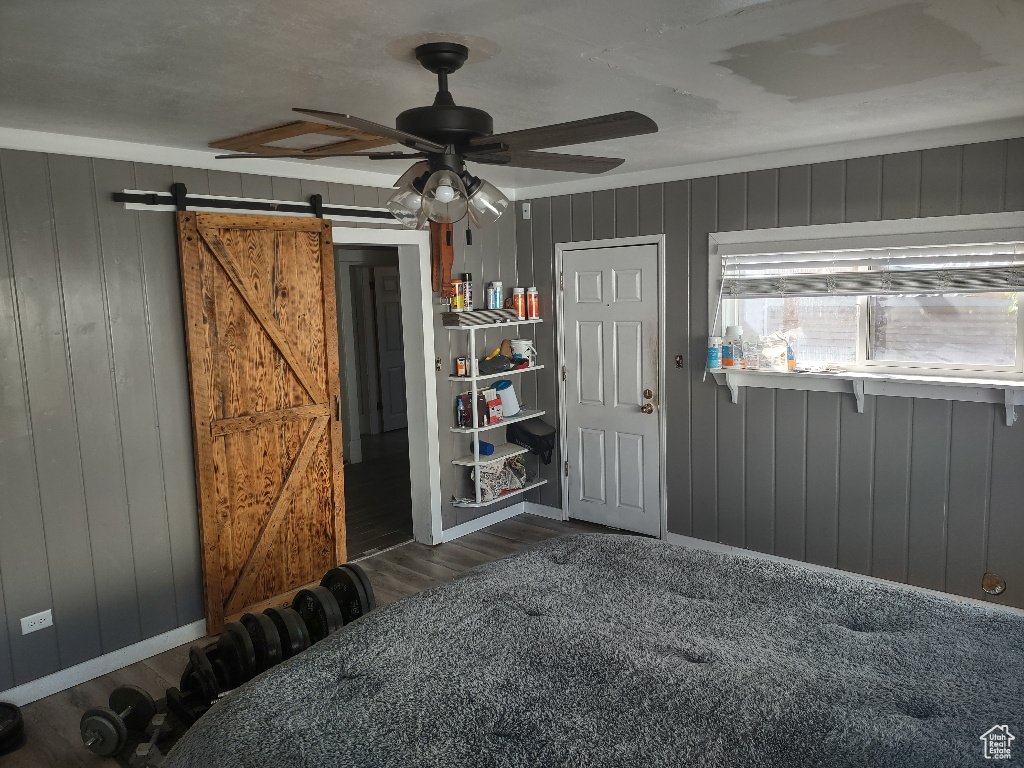 Unfurnished bedroom with a barn door, wooden walls, dark wood-type flooring, and ceiling fan