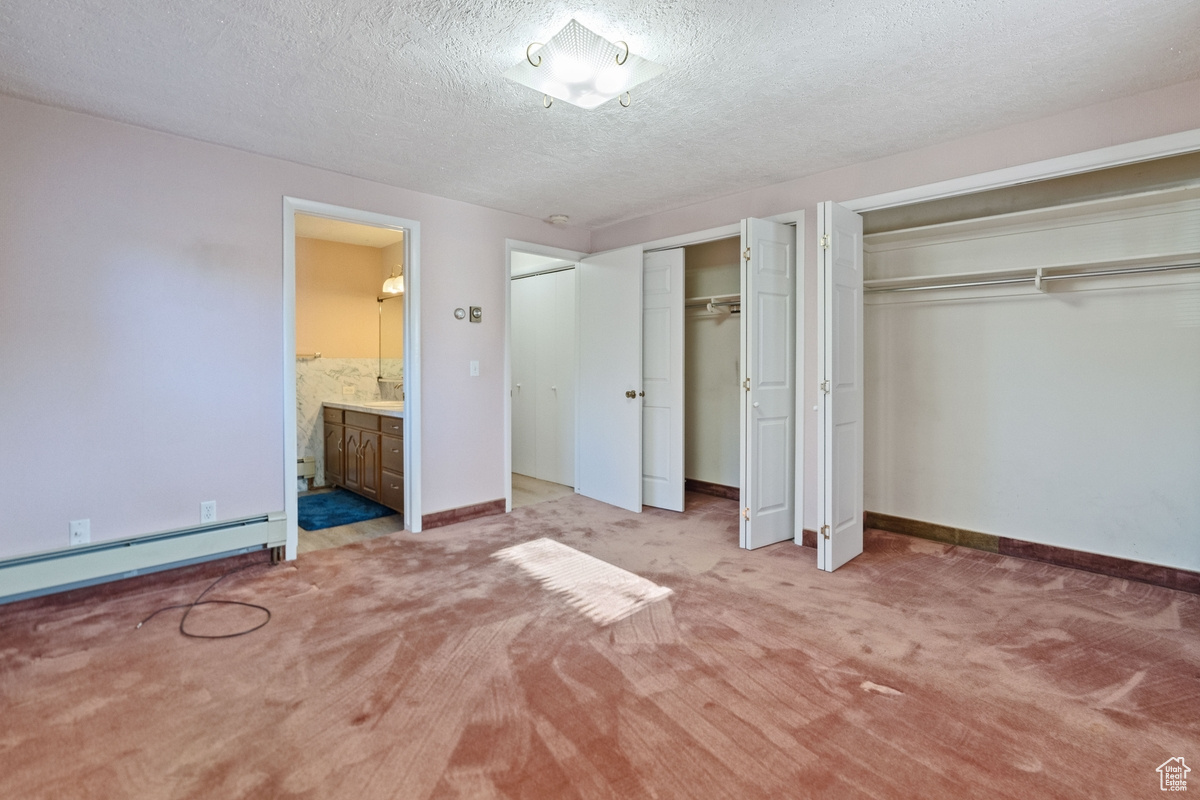 Unfurnished bedroom featuring connected bathroom, a textured ceiling, multiple closets, a baseboard heating unit, and light colored carpet