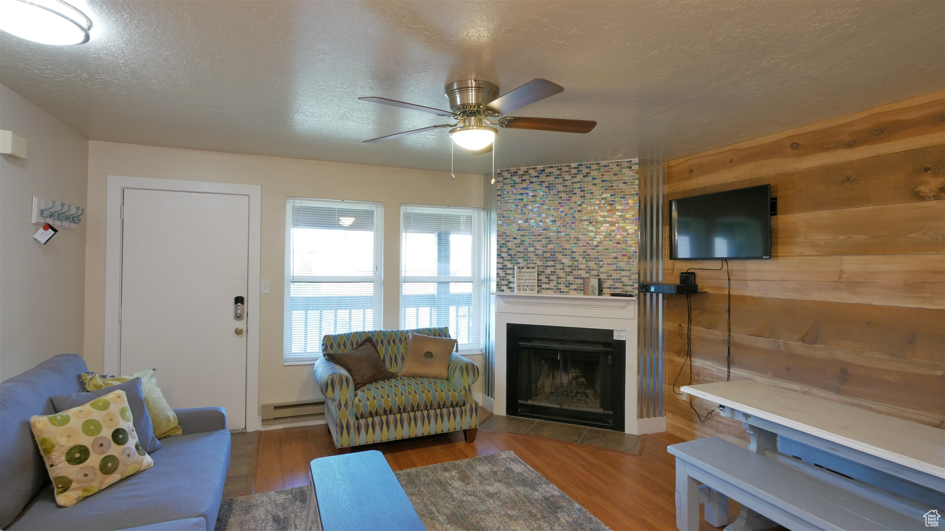 Living room with ceiling fan, wood walls, a textured ceiling, a baseboard heating unit, and hardwood / wood-style floors