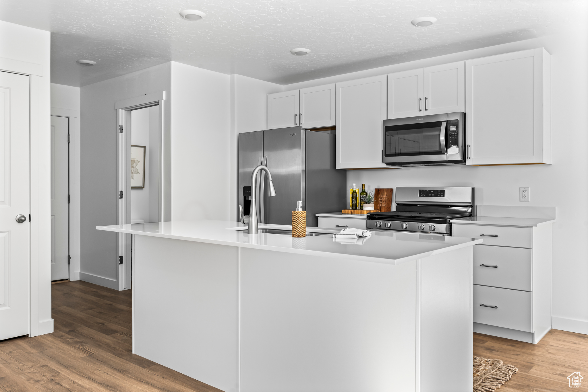 Kitchen featuring white cabinets, an island with sink, a textured ceiling, stainless steel appliances, and hardwood / wood-style floors