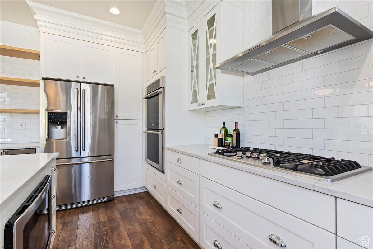 Kitchen featuring appliances with stainless steel finishes, dark wood-type flooring, tasteful backsplash, white cabinets, and wall chimney range hood