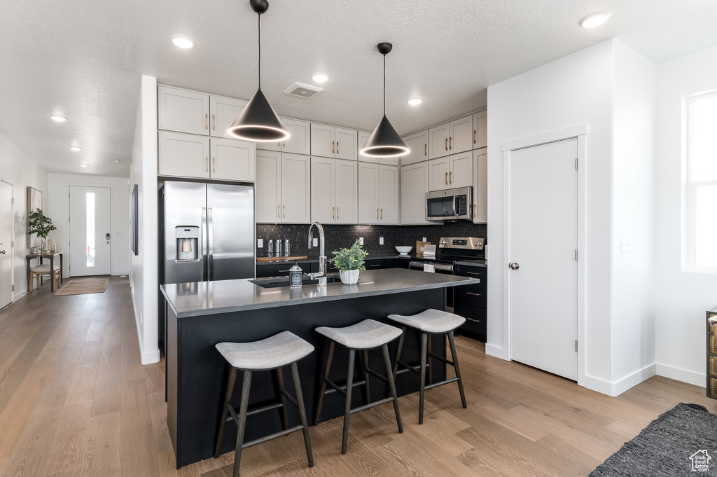 Kitchen featuring an island with sink, light wood-type flooring, stainless steel appliances, sink, and pendant lighting