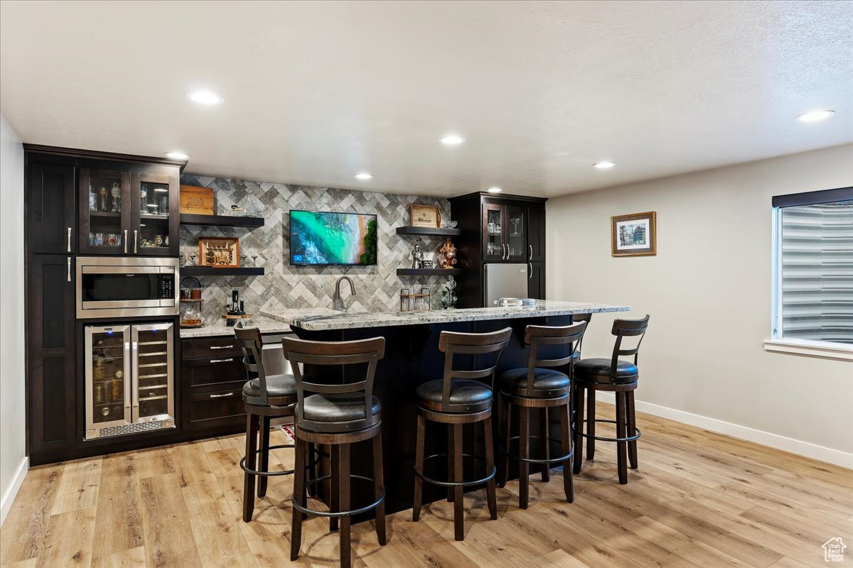 Bar featuring light wood-type flooring, light stone countertops, stainless steel microwave, wine cooler, and backsplash