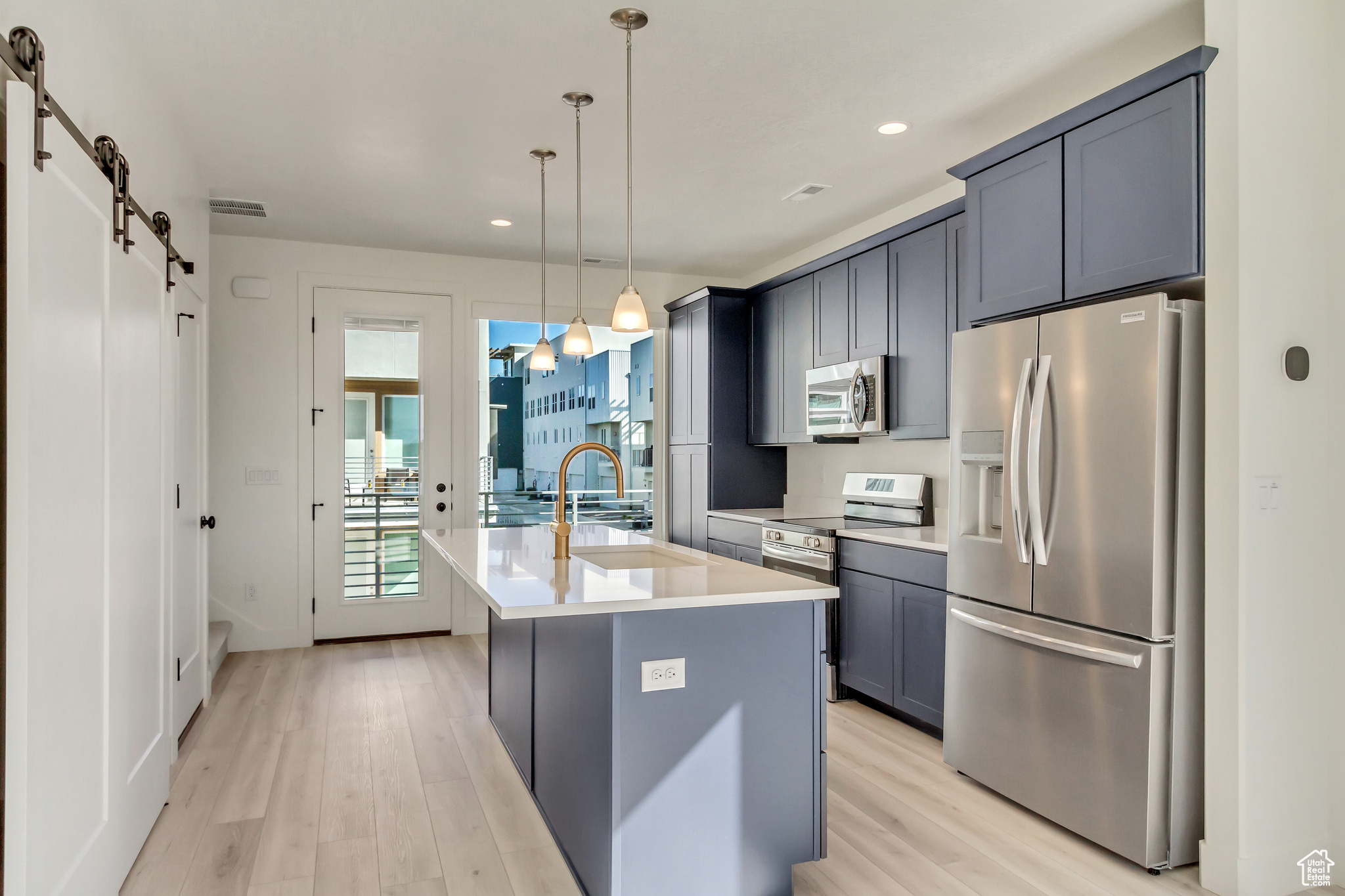 Kitchen with appliances with stainless steel finishes, a barn door, light hardwood / wood-style floors, pendant lighting, and a kitchen island with sink