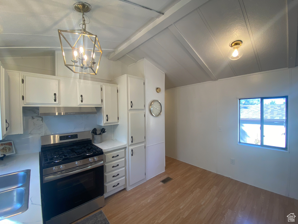 Kitchen with light wood-type flooring, white cabinets, stainless steel gas range oven, vaulted ceiling with beams, and decorative light fixtures