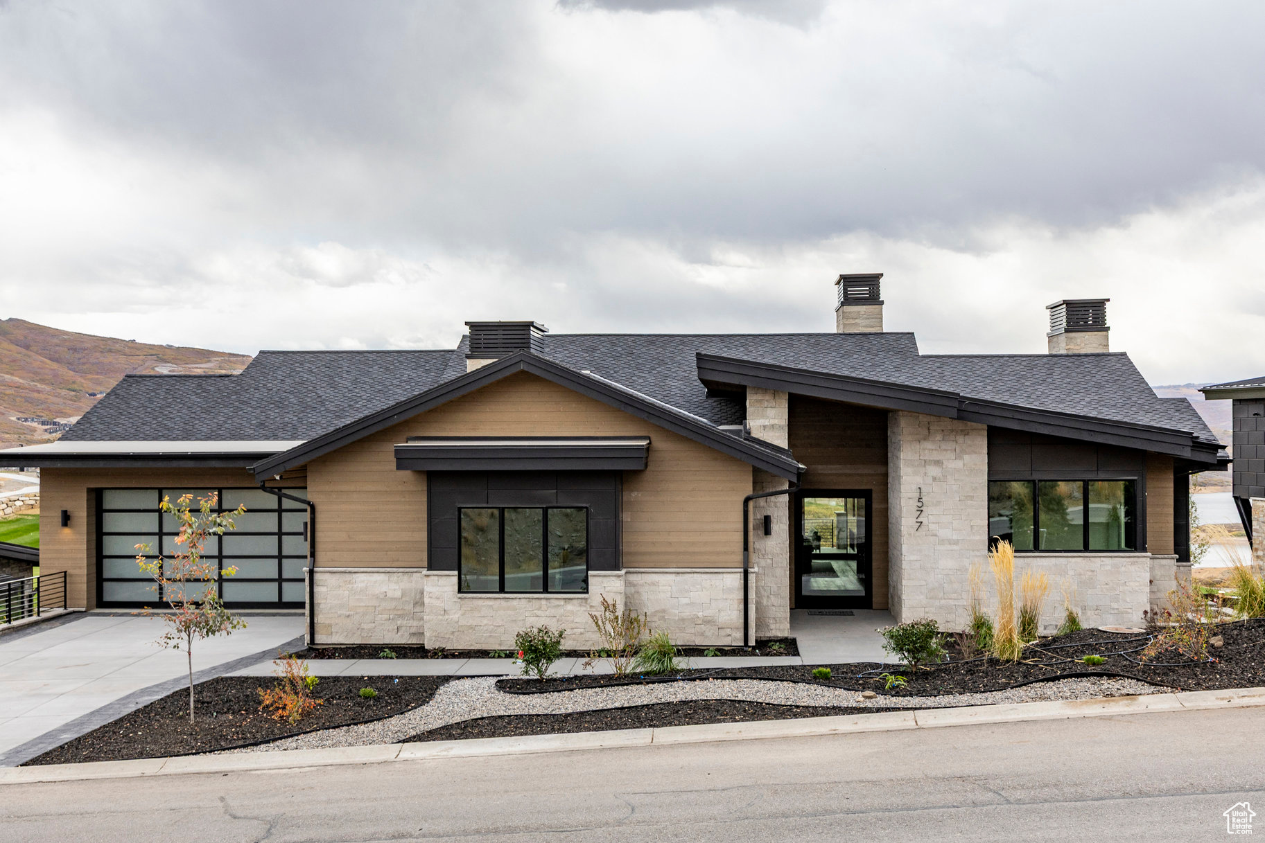 Modern stone-and-wood home with a dark roof, landscaped yard, and autumn hills behind it.