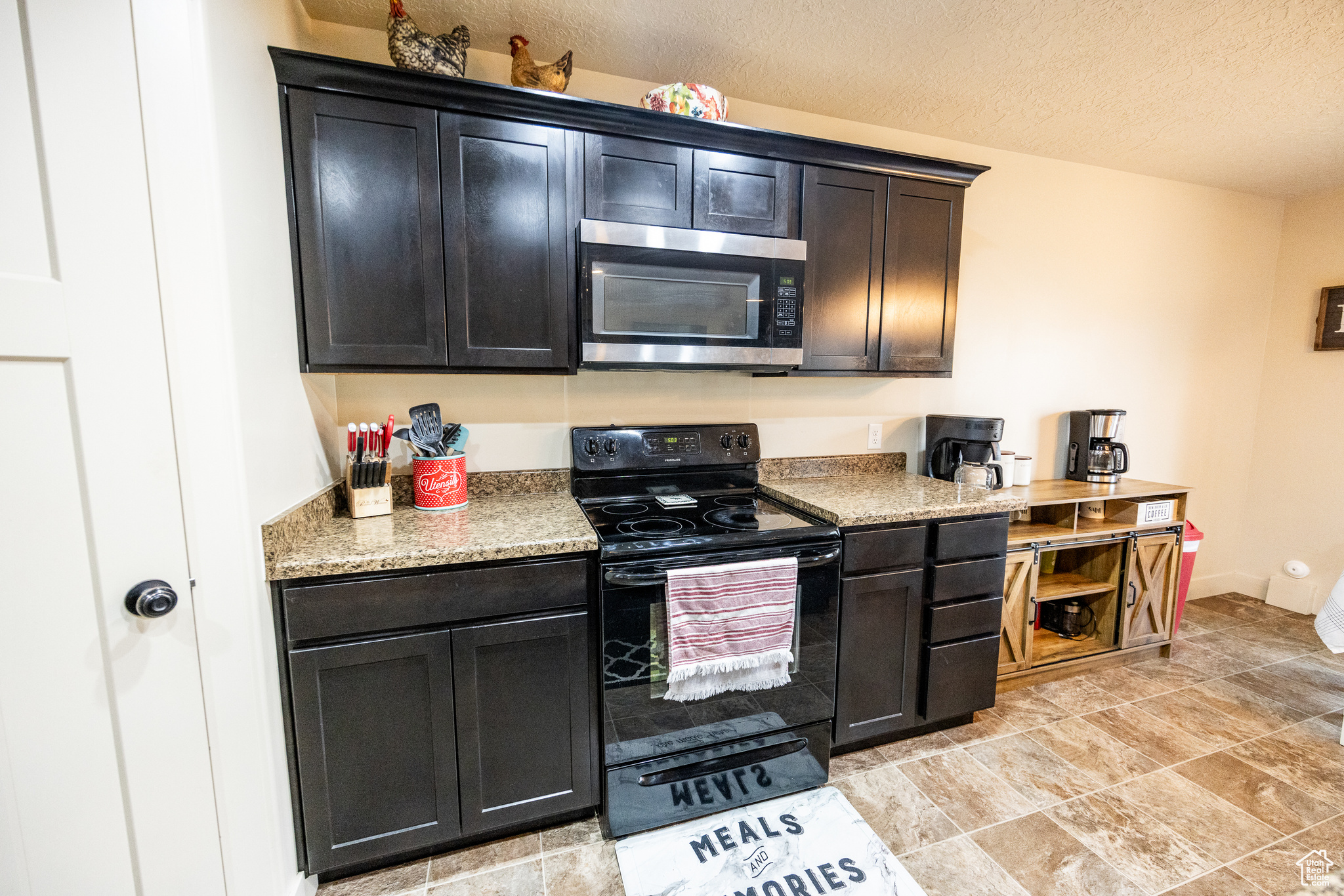 Kitchen featuring stainless steel microwave, dark cabinets, light stone countertops, a textured ceiling, and black electric range