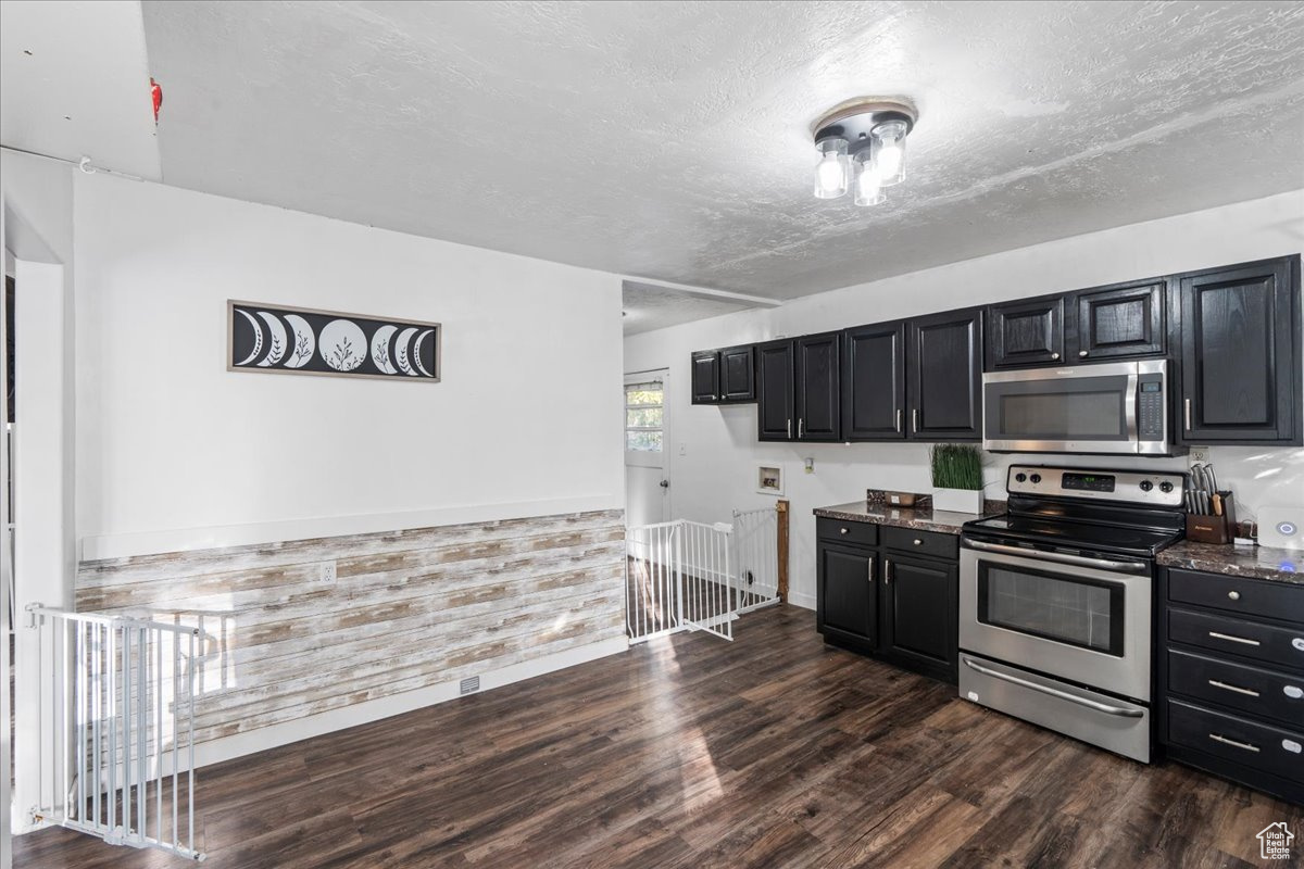 Kitchen with a textured ceiling, dark stone countertops, dark wood-type flooring, wooden walls, and stainless steel appliances