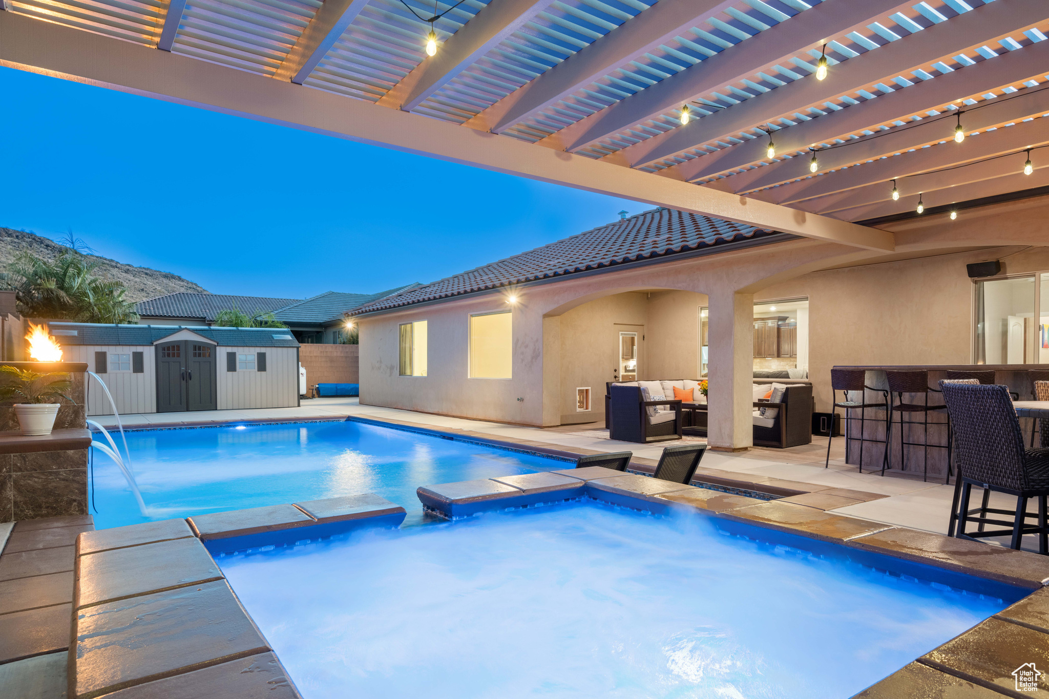 Pool at dusk featuring a bar, a pergola, pool water feature, a storage shed, and a patio