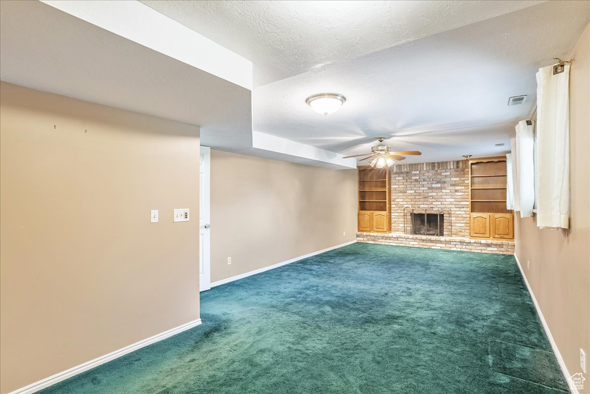 Unfurnished living room featuring a textured ceiling, a brick fireplace, ceiling fan, carpet, and built in shelves