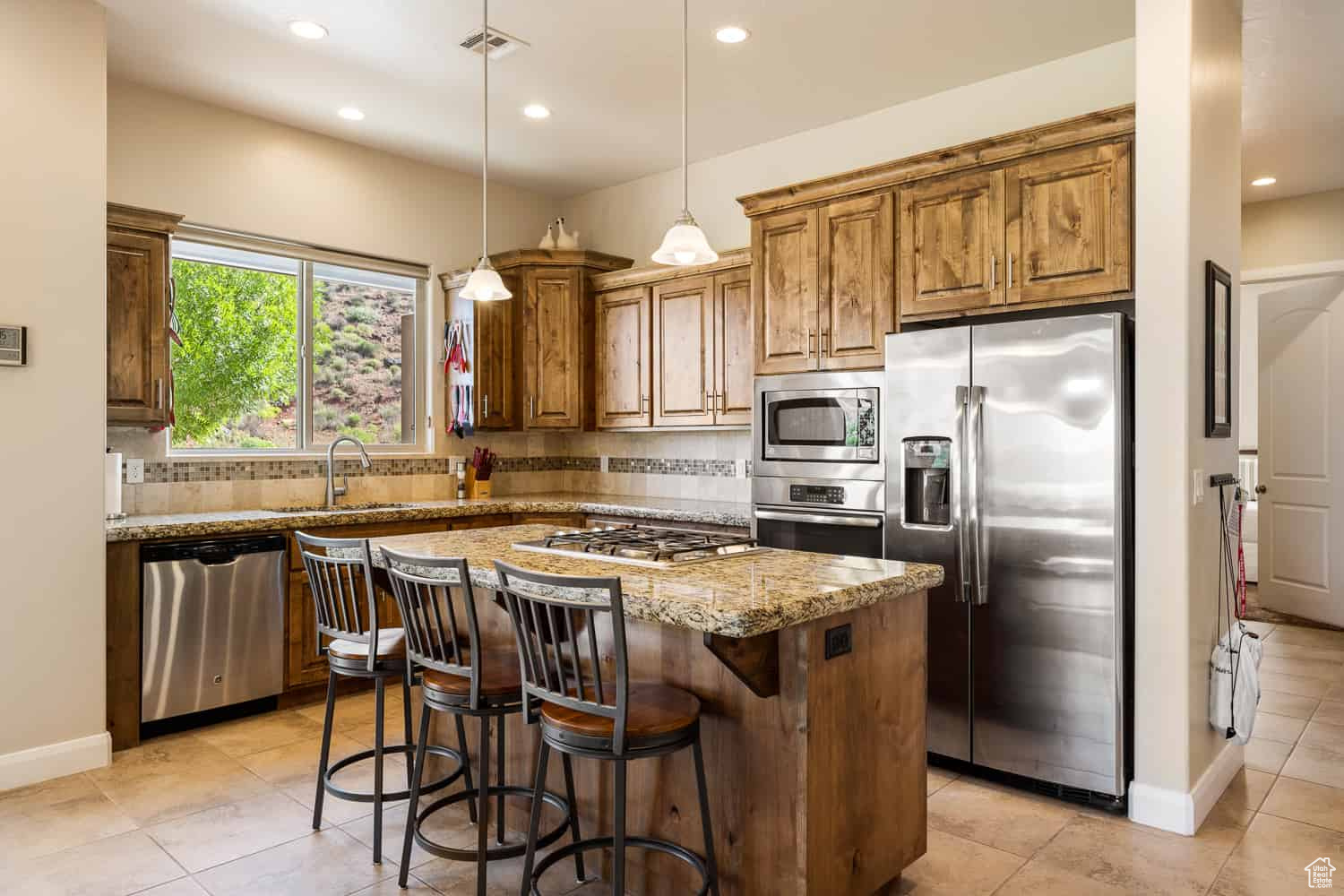 Kitchen with light stone counters, light tile patterned floors, appliances with stainless steel finishes, a center island, and decorative light fixtures