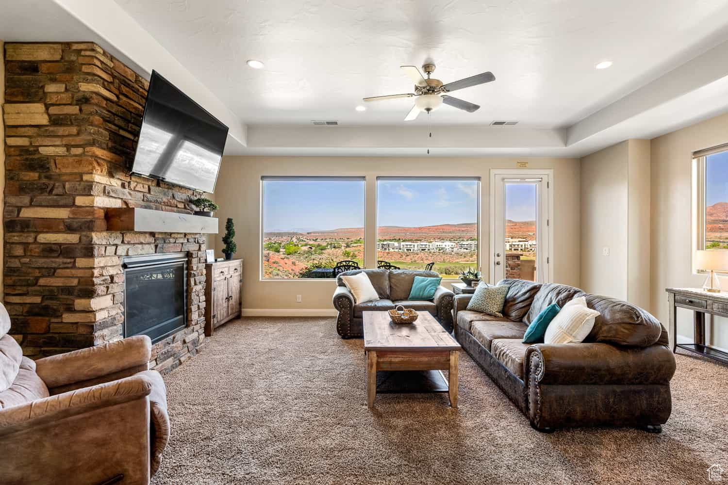 Carpeted living room featuring a stone fireplace, a healthy amount of sunlight, a raised ceiling, and ceiling fan