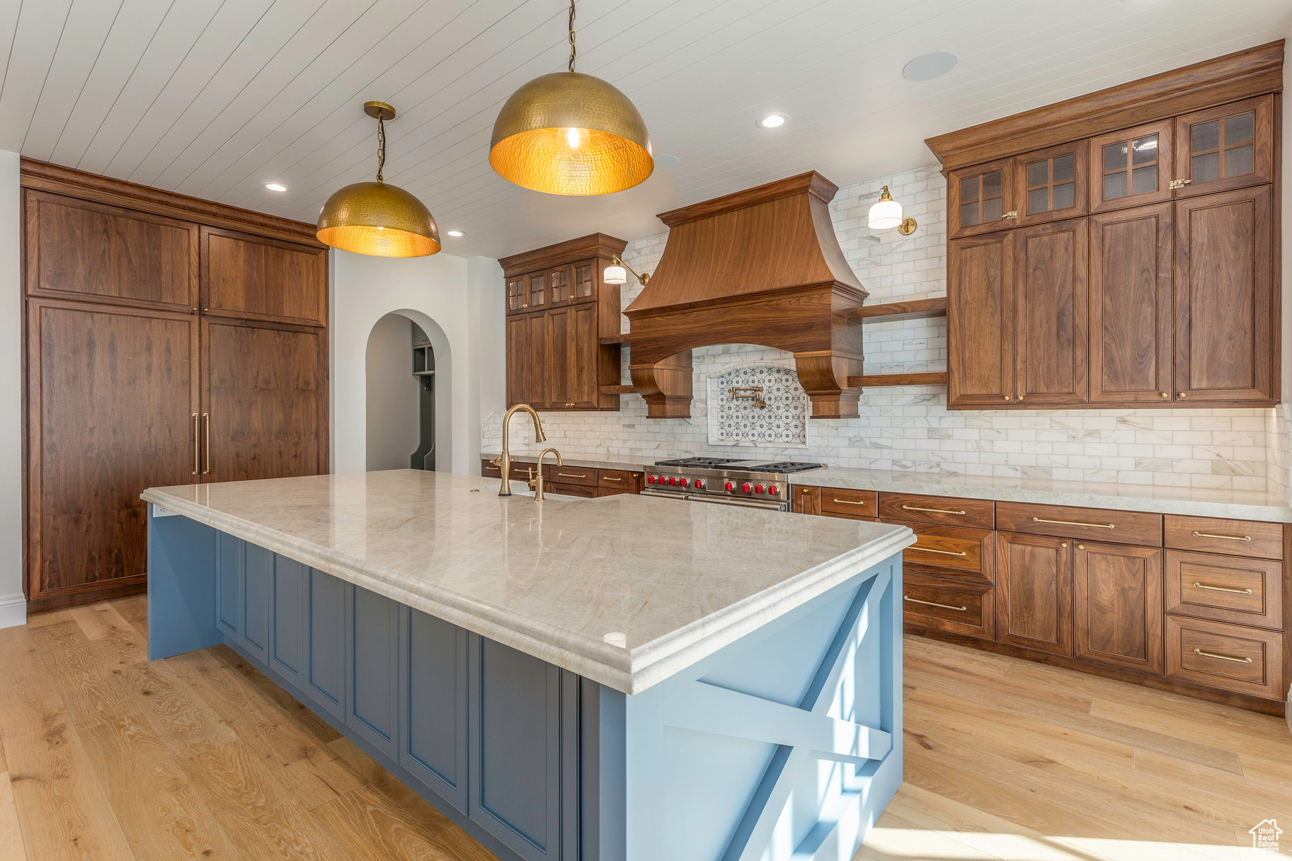 Kitchen with premium range hood, light stone counters, decorative light fixtures, light hardwood / wood-style floors, and an island with sink