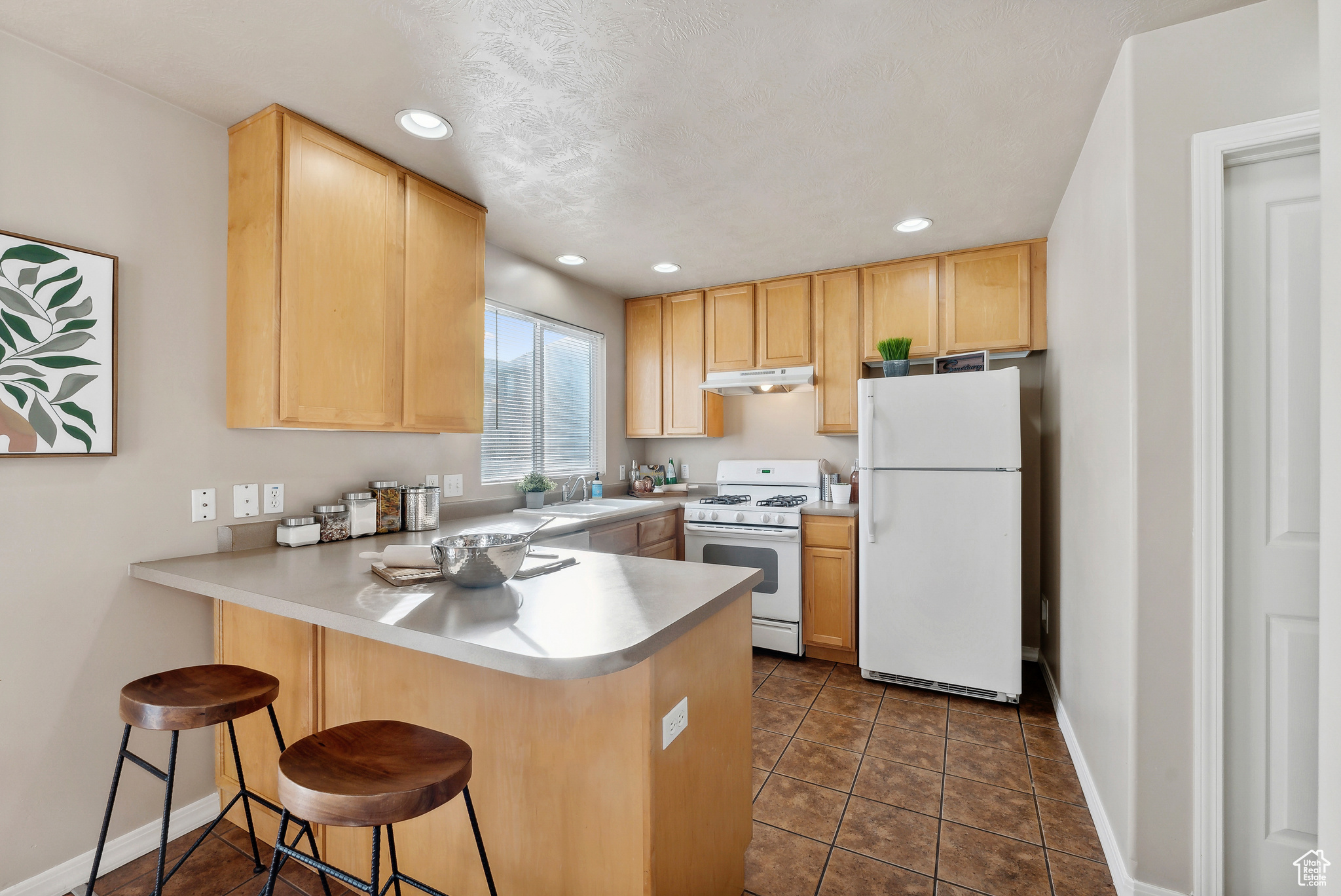 Kitchen featuring kitchen peninsula, light brown cabinetry, a kitchen breakfast bar, white appliances, and dark tile patterned floors