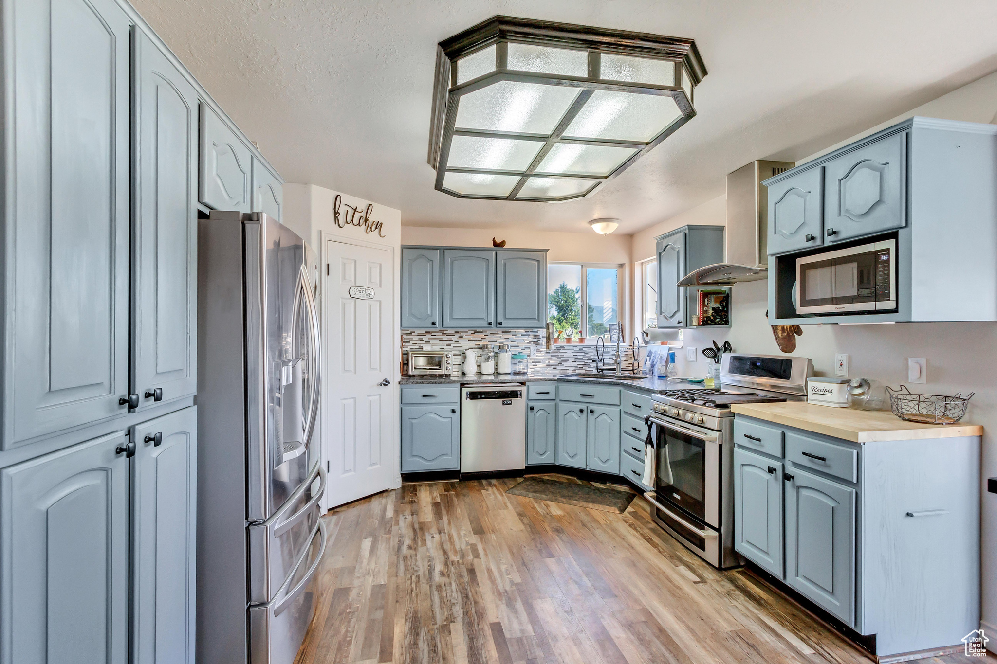 Kitchen featuring sink, wall chimney exhaust hood, light wood-type flooring, appliances with stainless steel finishes, and tasteful backsplash