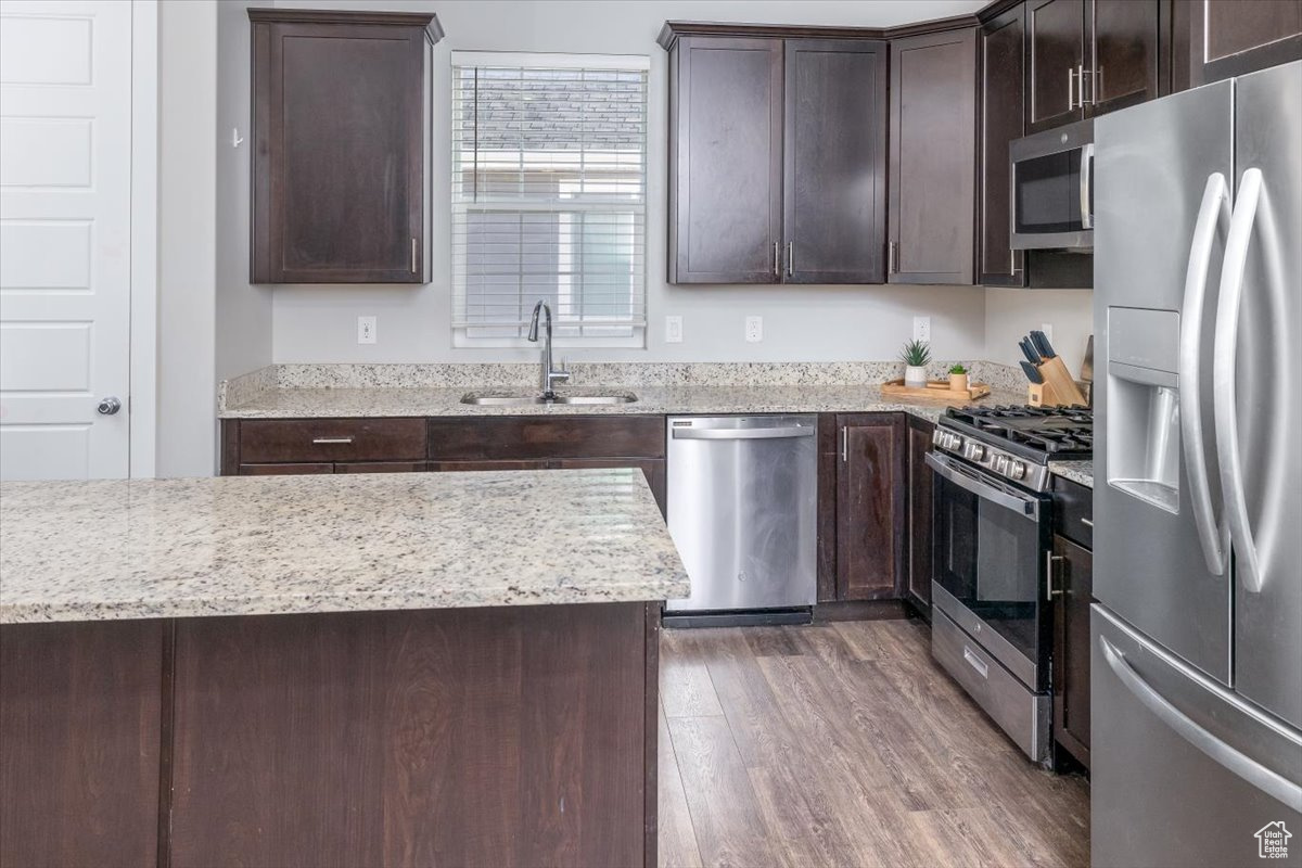 Kitchen with appliances with stainless steel finishes, light stone counters, dark brown cabinets, dark wood-type flooring, and sink