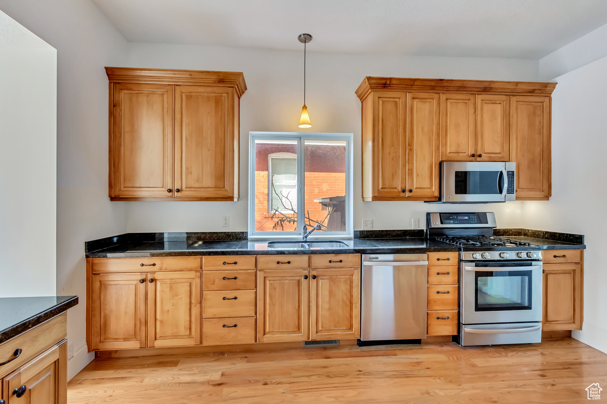 Kitchen with appliances with stainless steel finishes, dark stone counters, sink, decorative light fixtures, and light hardwood / wood-style flooring