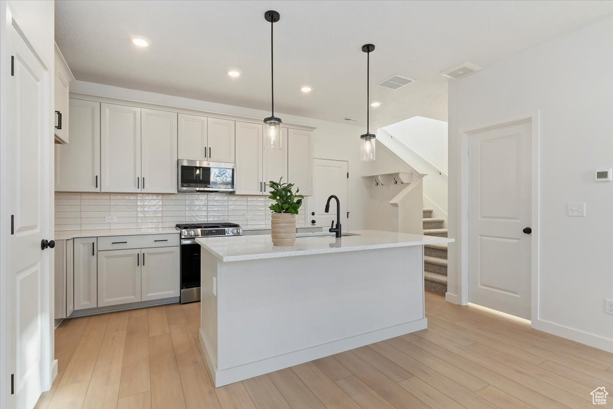 Kitchen with appliances with stainless steel finishes, light wood-type flooring, decorative light fixtures, white cabinetry, and an island with sink