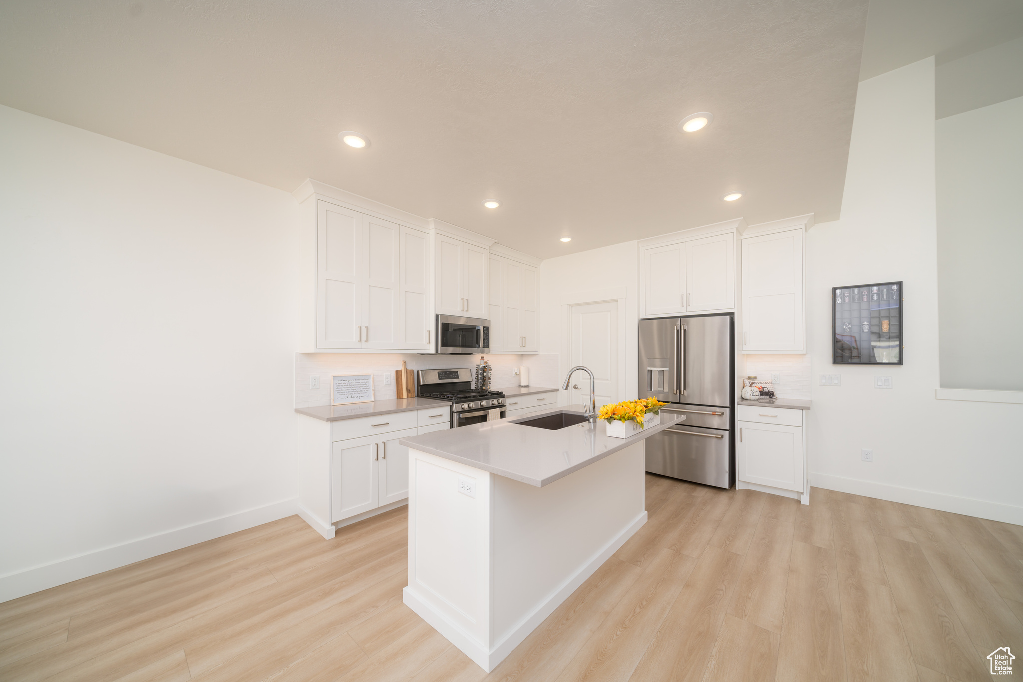 Kitchen with light wood-type flooring, stainless steel appliances, sink, white cabinets, and an island with sink