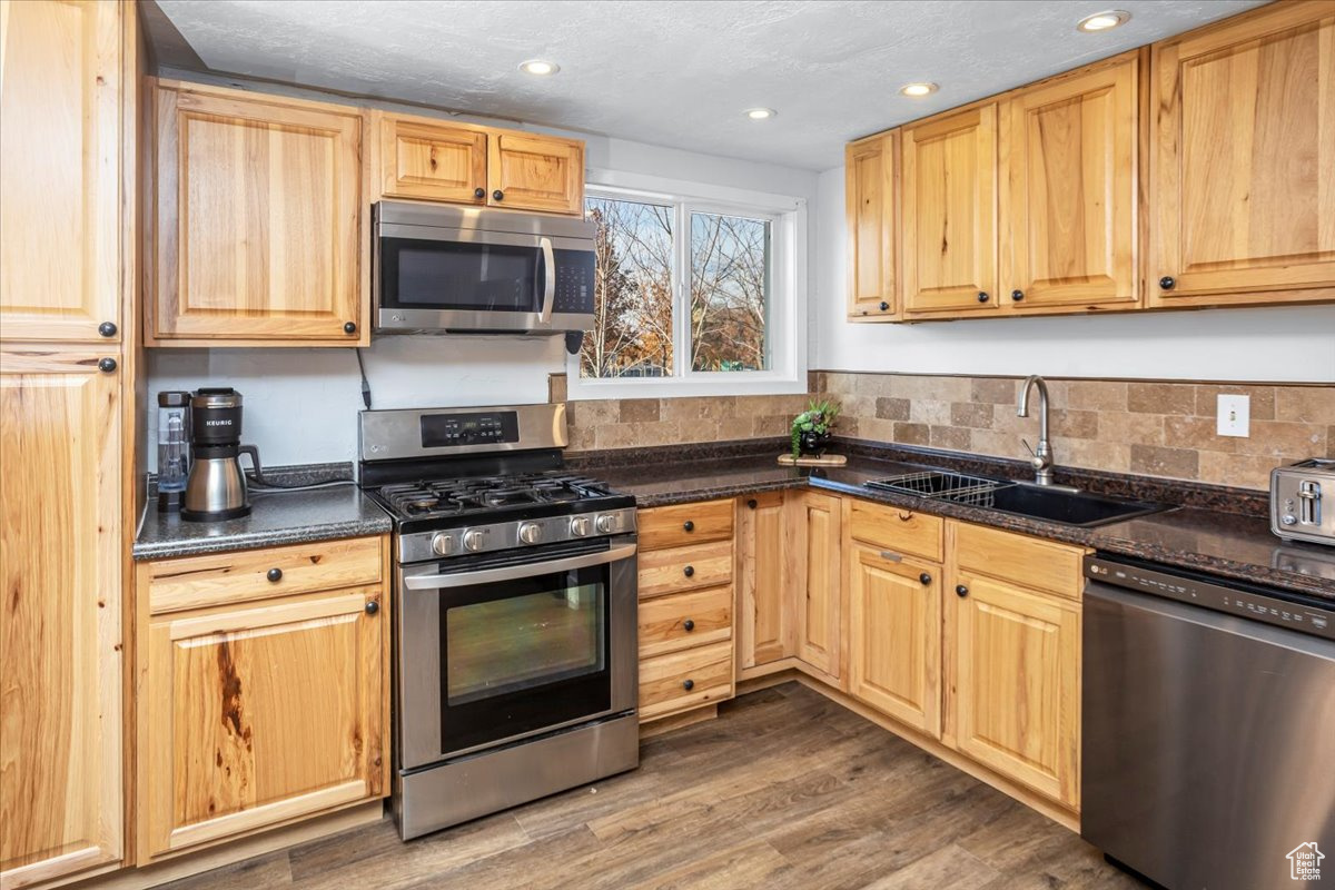 Kitchen with sink, dark wood-type flooring, dark stone countertops, light brown cabinetry, and appliances with stainless steel finishes