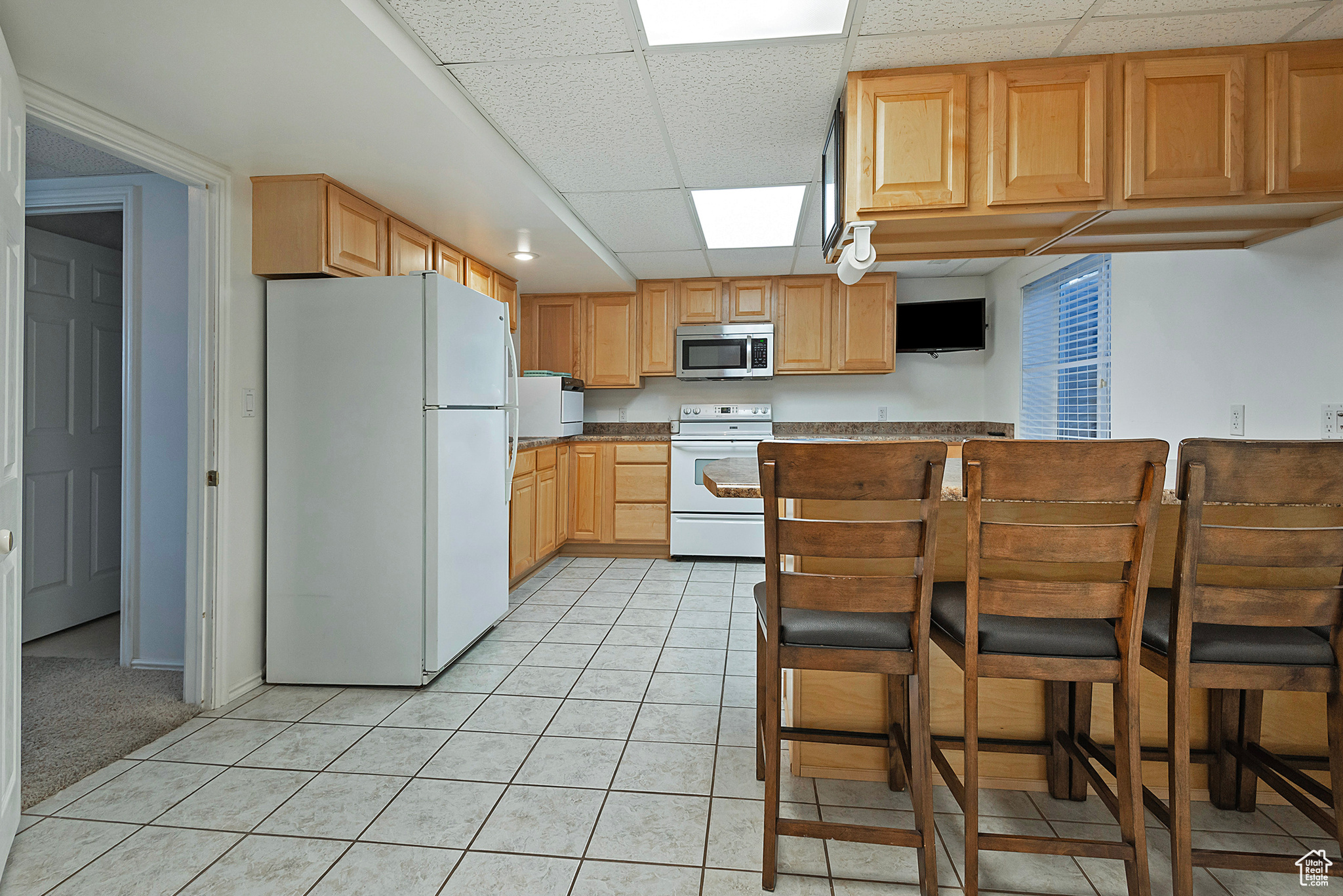 Kitchen with a paneled ceiling, light brown cabinets, white appliances, and light tile patterned floors