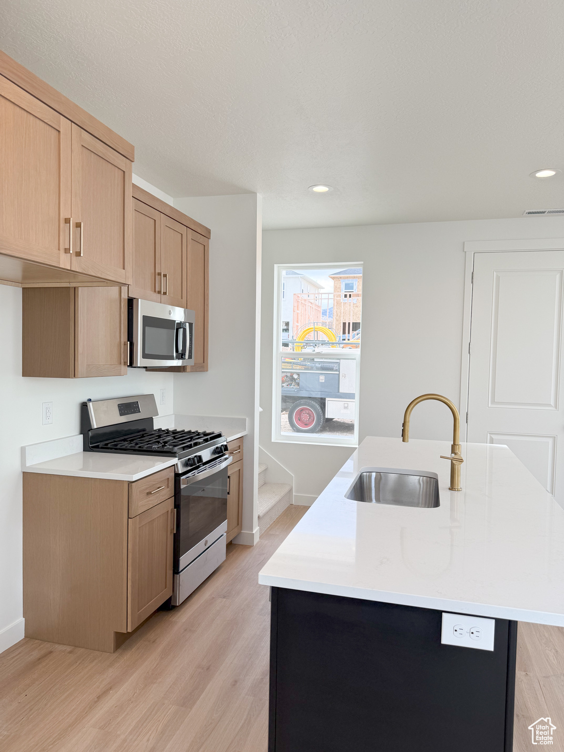 Kitchen with appliances with stainless steel finishes, light wood-type flooring, sink, light brown cabinets, and a center island with sink