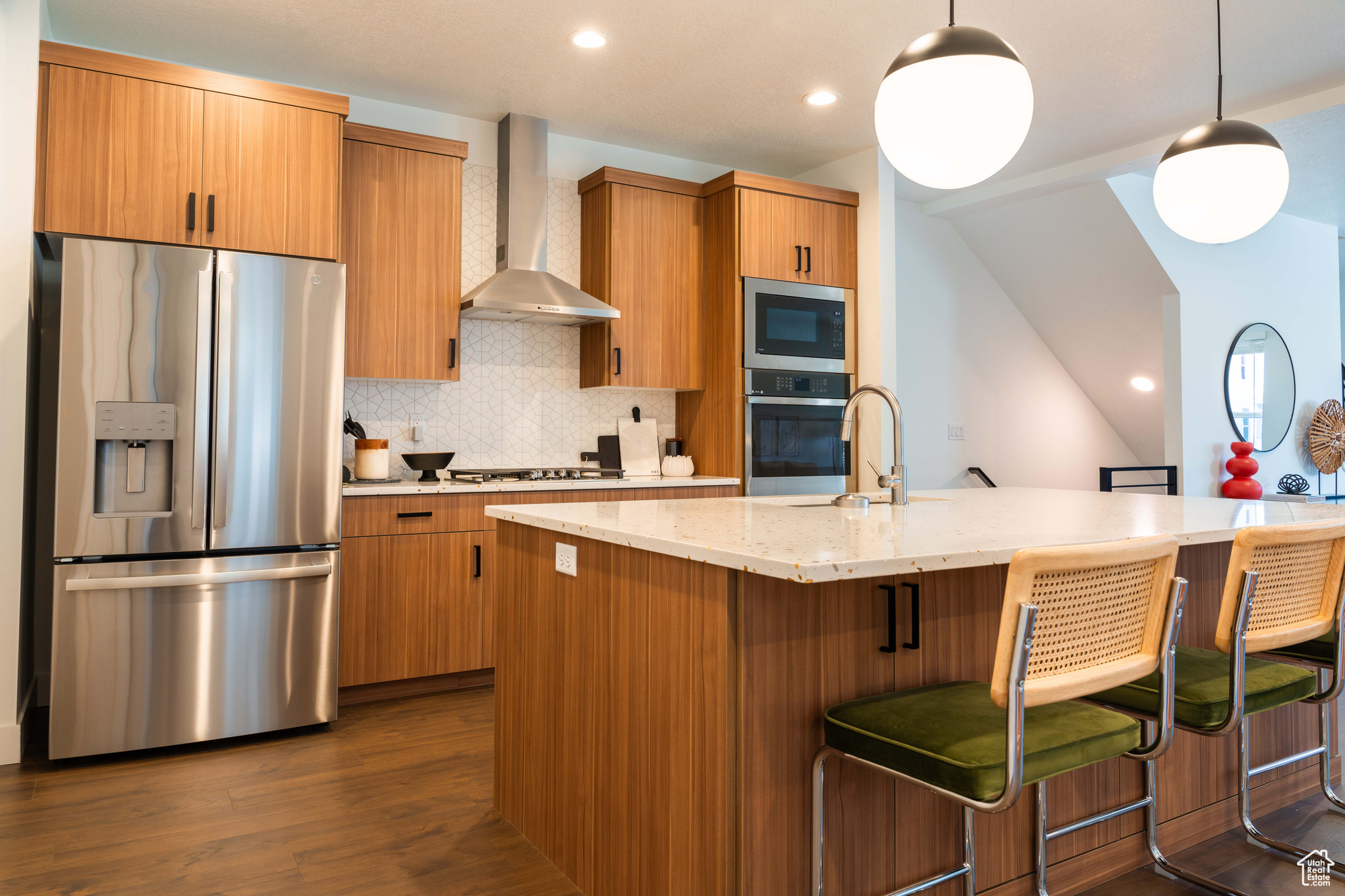 Kitchen with a center island with sink, wall chimney range hood, appliances with stainless steel finishes, decorative light fixtures, and dark hardwood / wood-style flooring