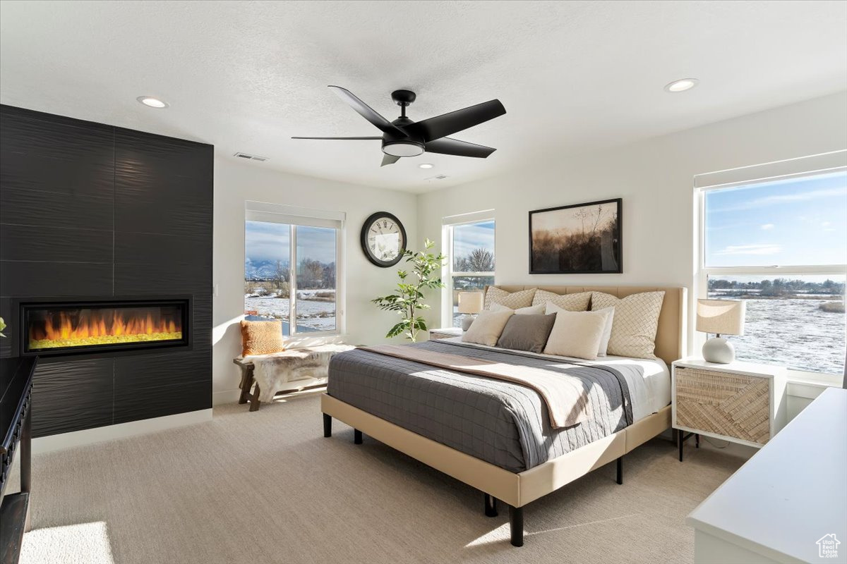 Bedroom featuring multiple windows, ceiling fan, a large fireplace, and light colored carpet