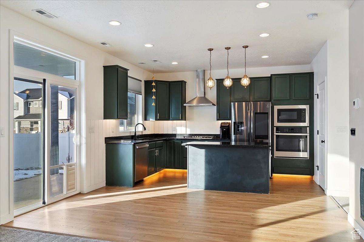 Kitchen featuring a center island, wall chimney exhaust hood, decorative light fixtures, appliances with stainless steel finishes, and light wood-type flooring